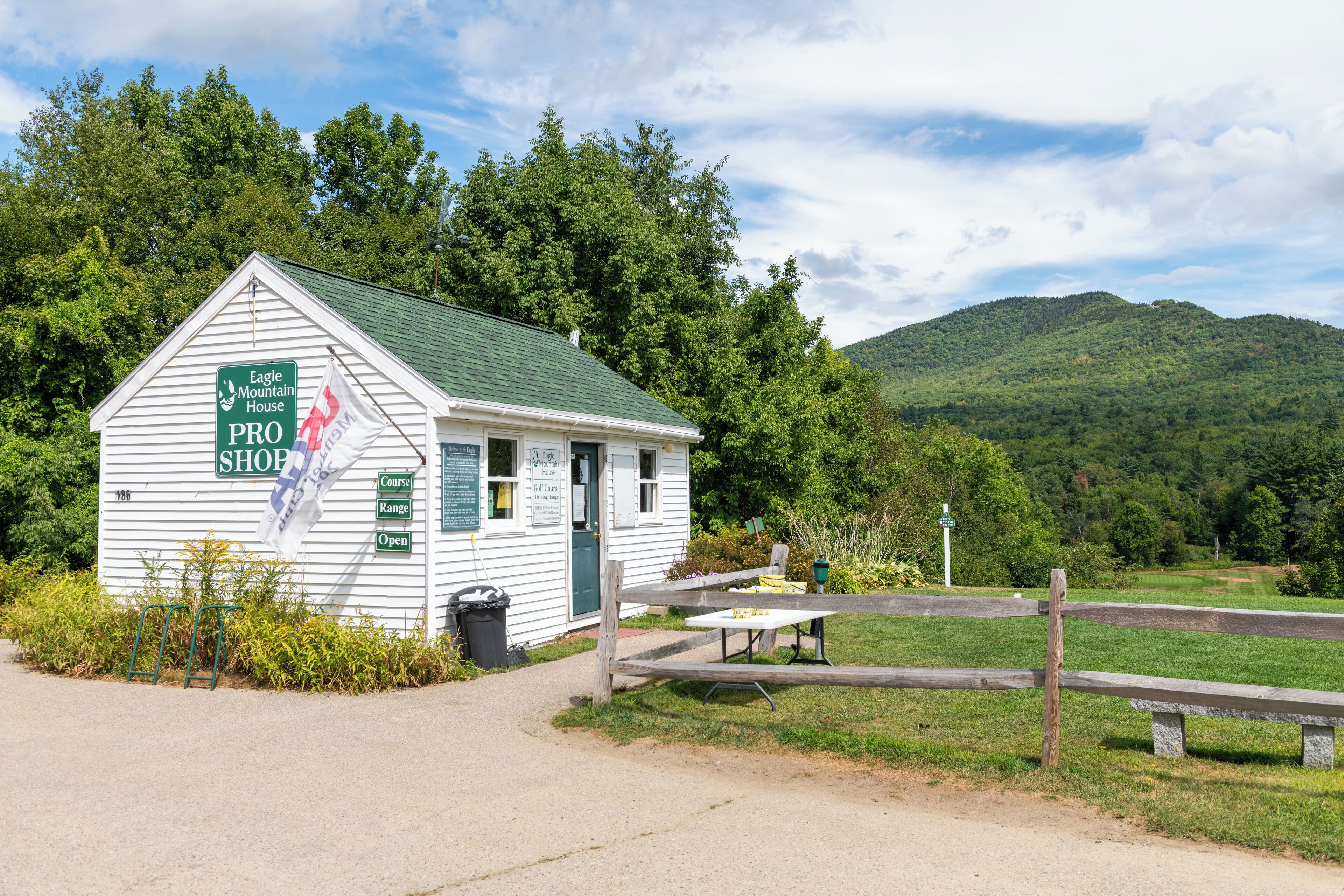 Un pequeño edificio blanco con techo verde, identificado como 'Eagle Mountain House PRO Shop', ubicado en un paisaje verde exuberante con una montaña al fondo.