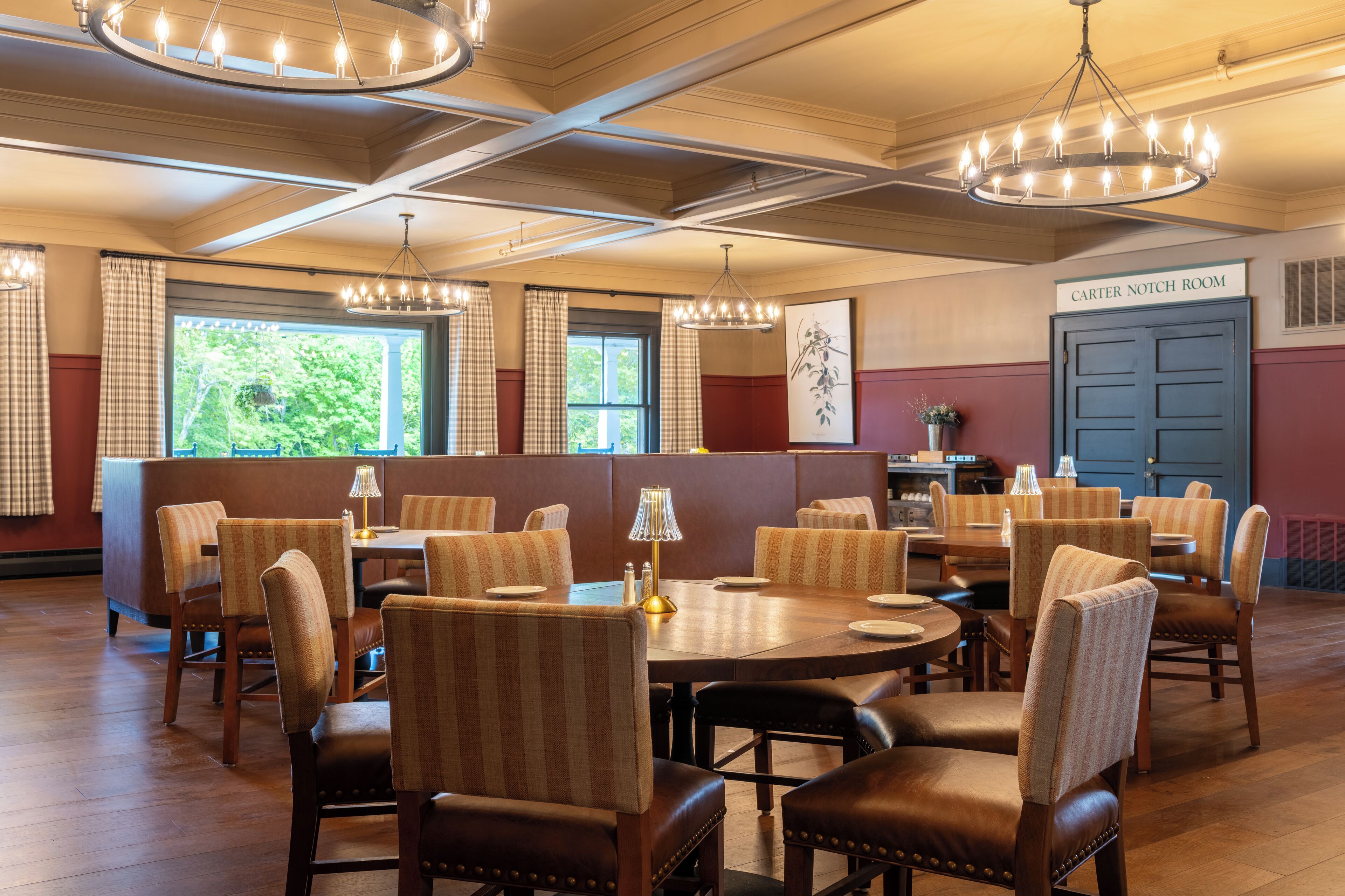 A dining area with multiple tables and chairs, a chandelier, and a view of greenery outside.