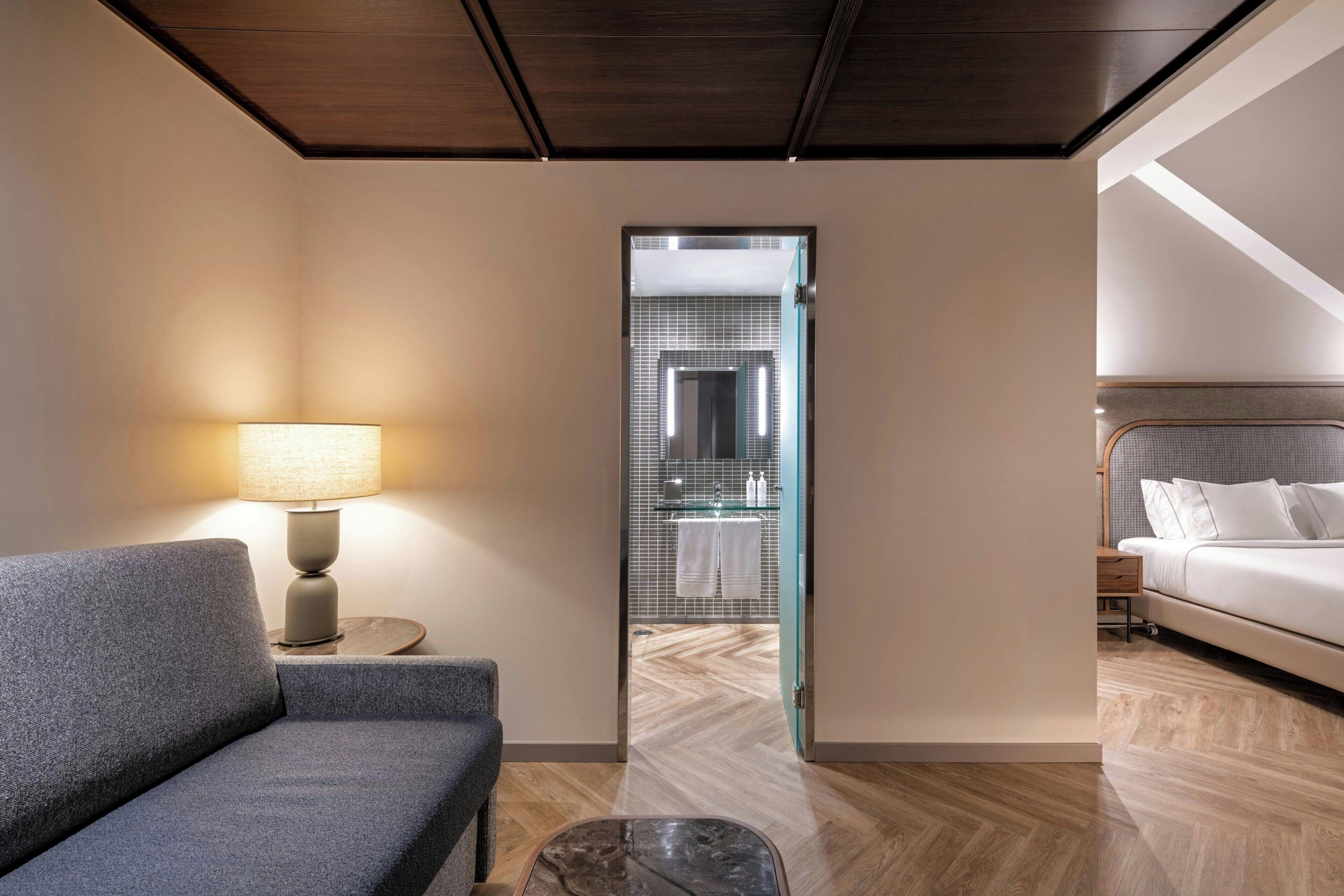 Habitación de hotel con sofá, lámpara, una mesita, cama y baño visible a través de una puerta de cristal.