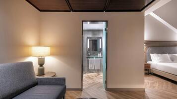 Habitación de hotel con sofá, lámpara, una mesita, cama y baño visible a través de una puerta de cristal.