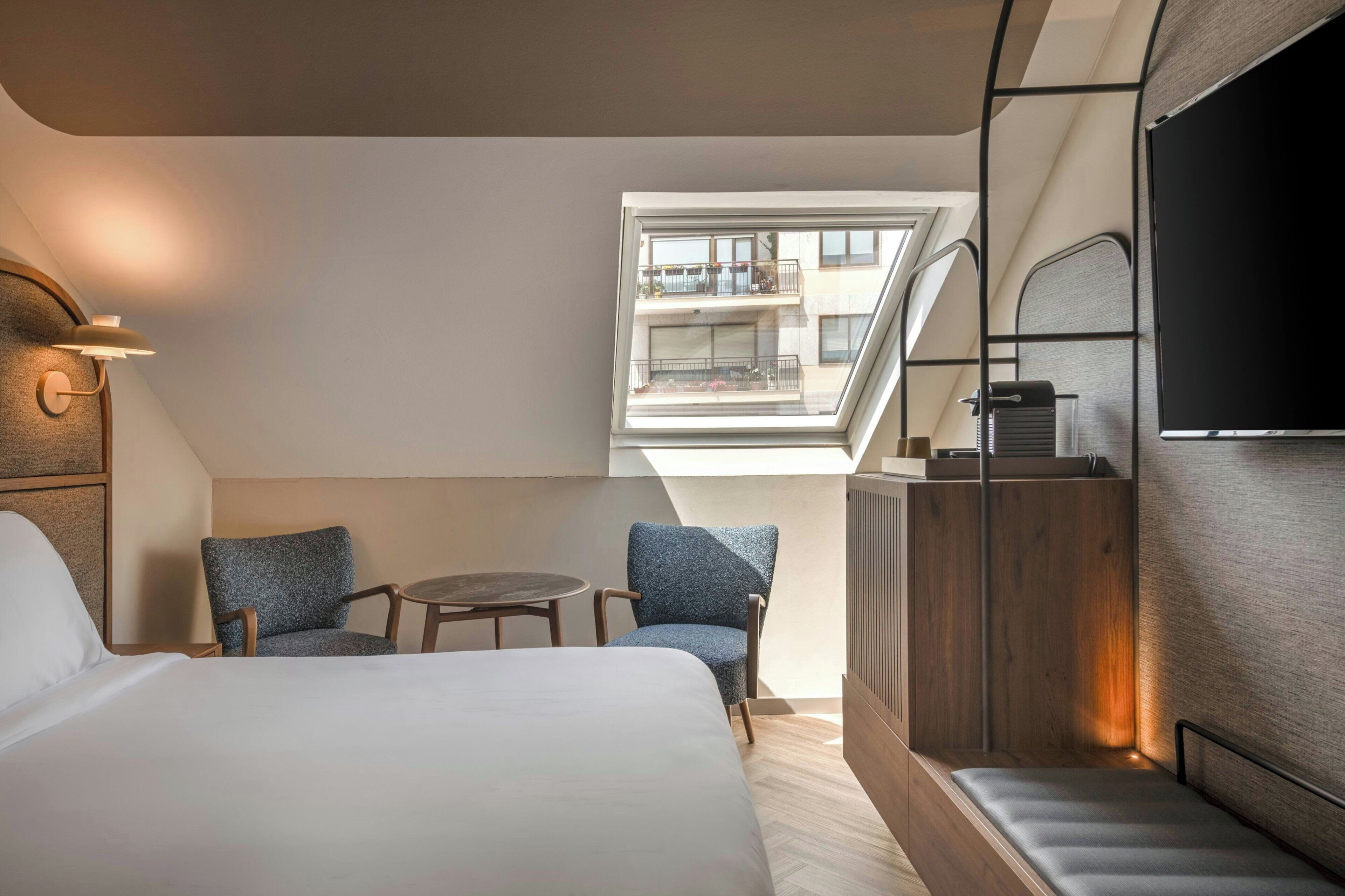 A hotel room with a bed, two chairs, a small table, a TV, and a balcony with a view.