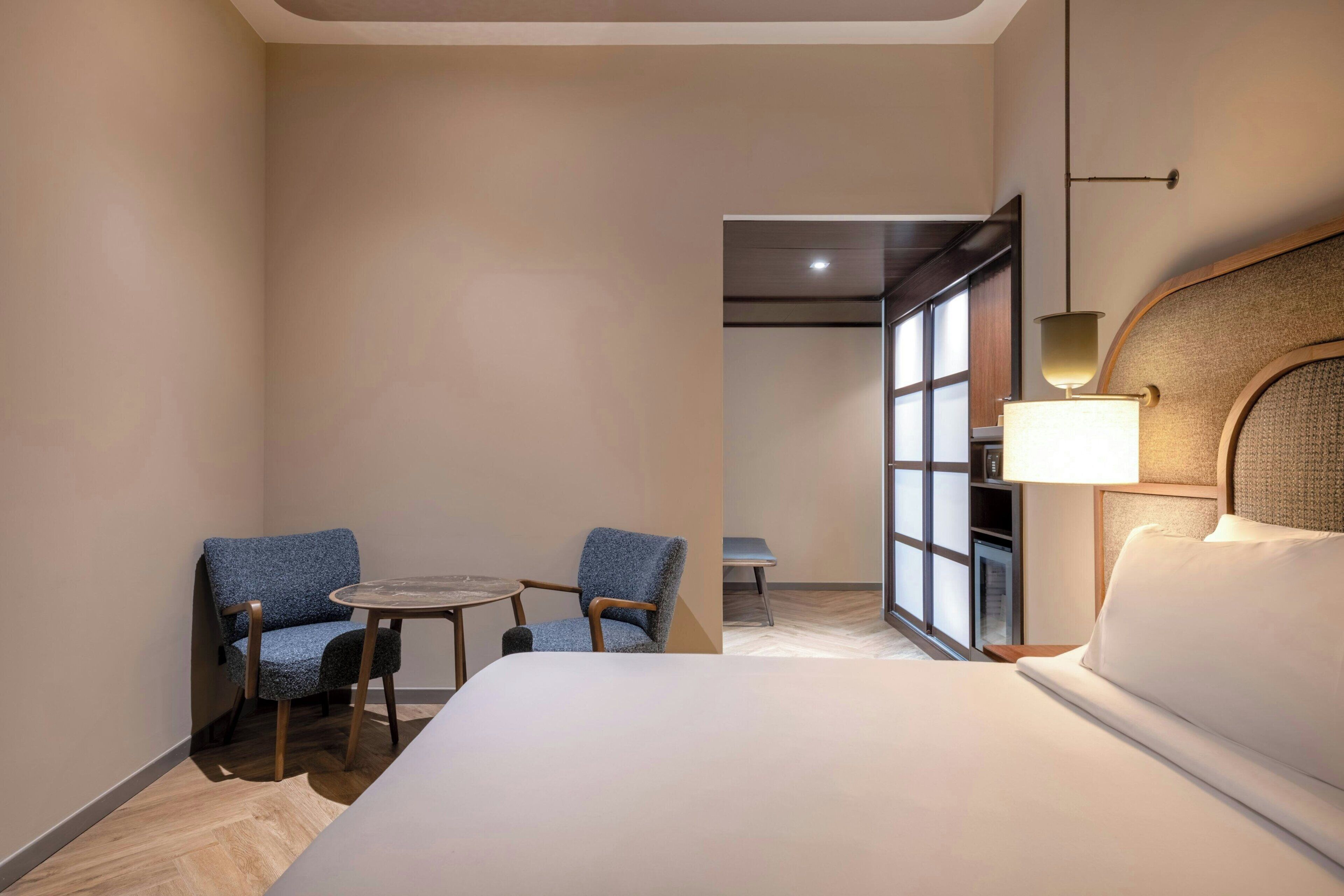 A hotel room with a bed, two chairs, a small table, a lamp, and a view of the outside through a window.