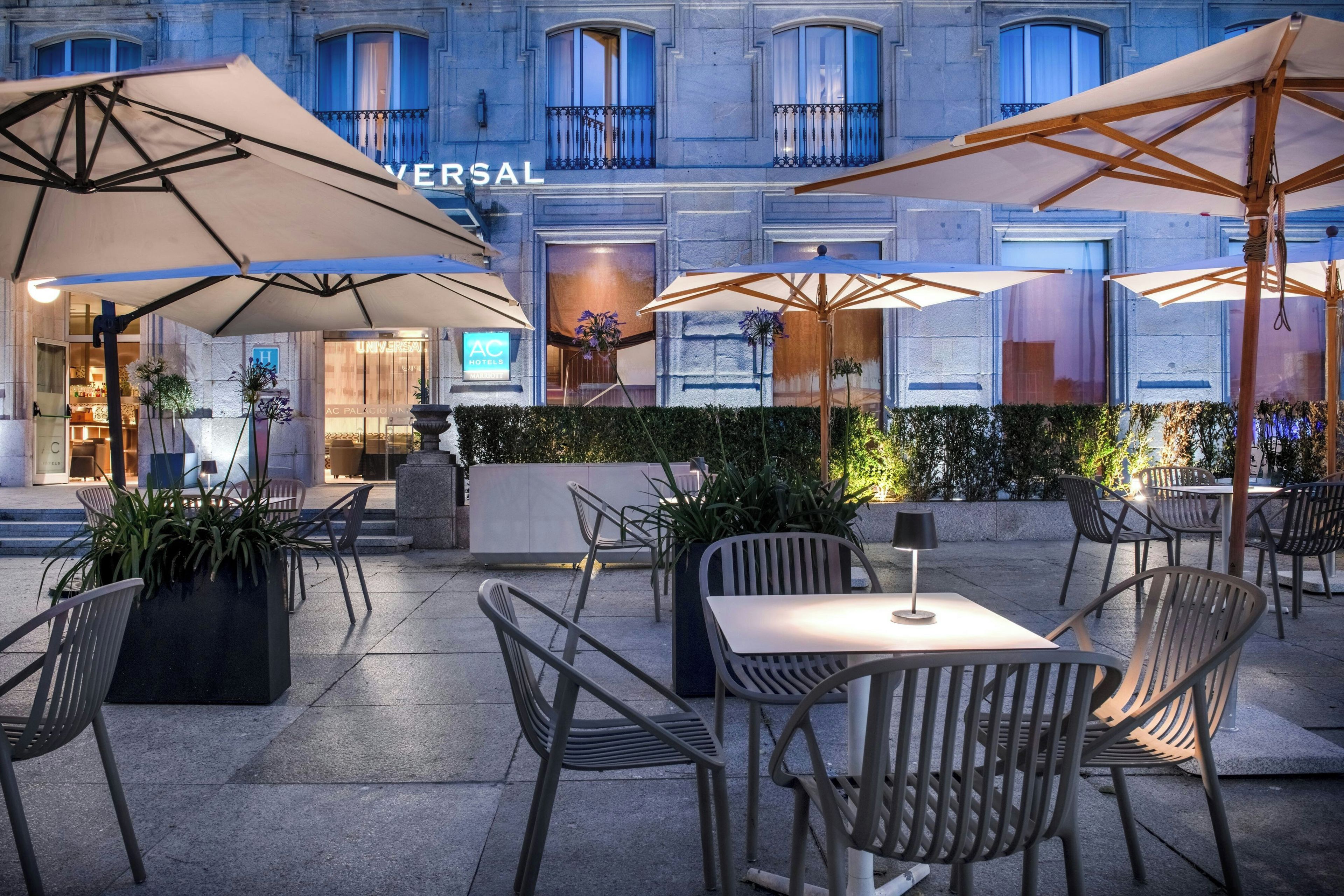 Un área de estar al aire libre con mesas y sillas, sombrillas y un edificio con letrero 'UNIVERSAL' al fondo.