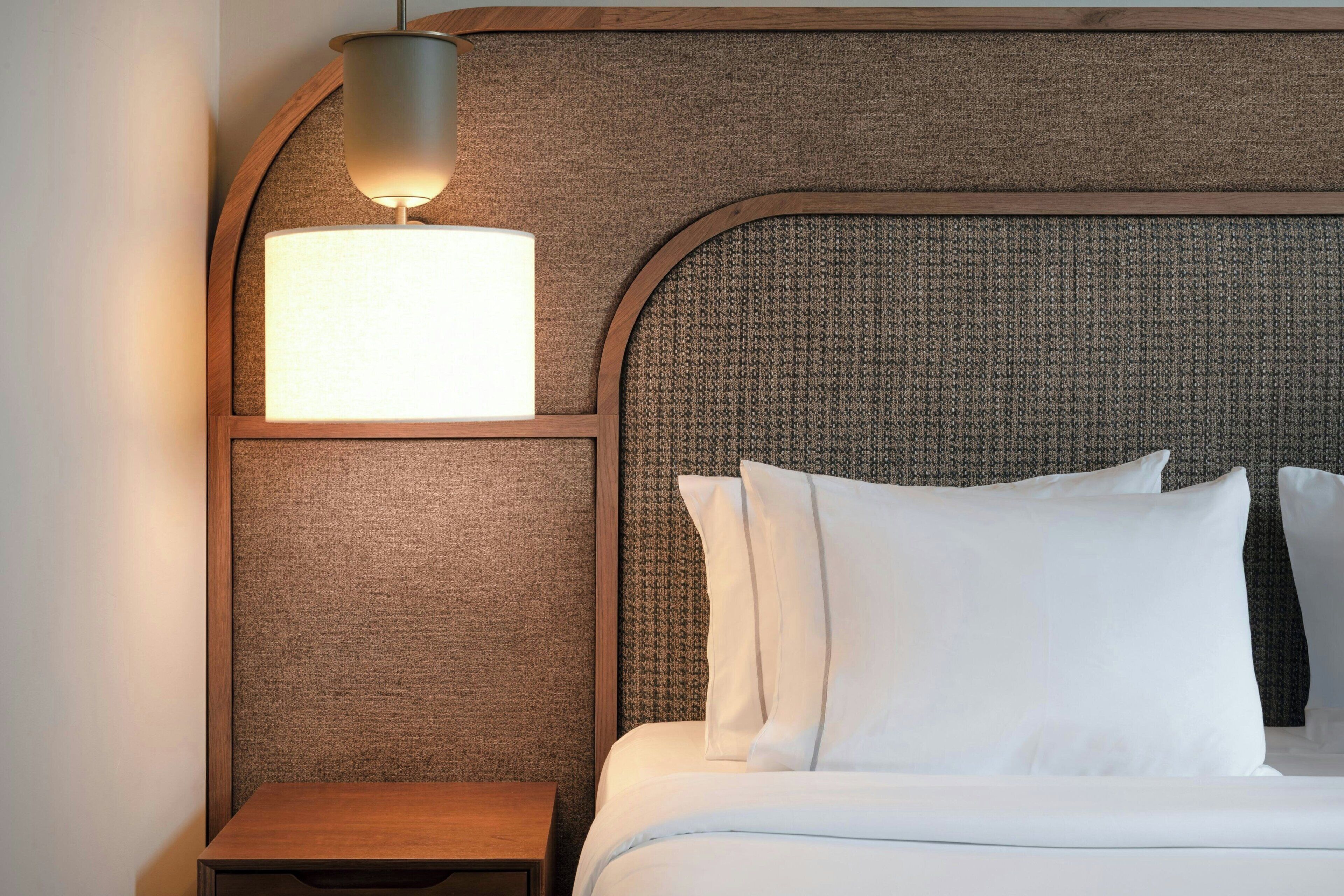 Un lit bien fait, avec du linge de lit blanc et des oreillers, une petite table de chevet en bois et une lampe fixée au mur.