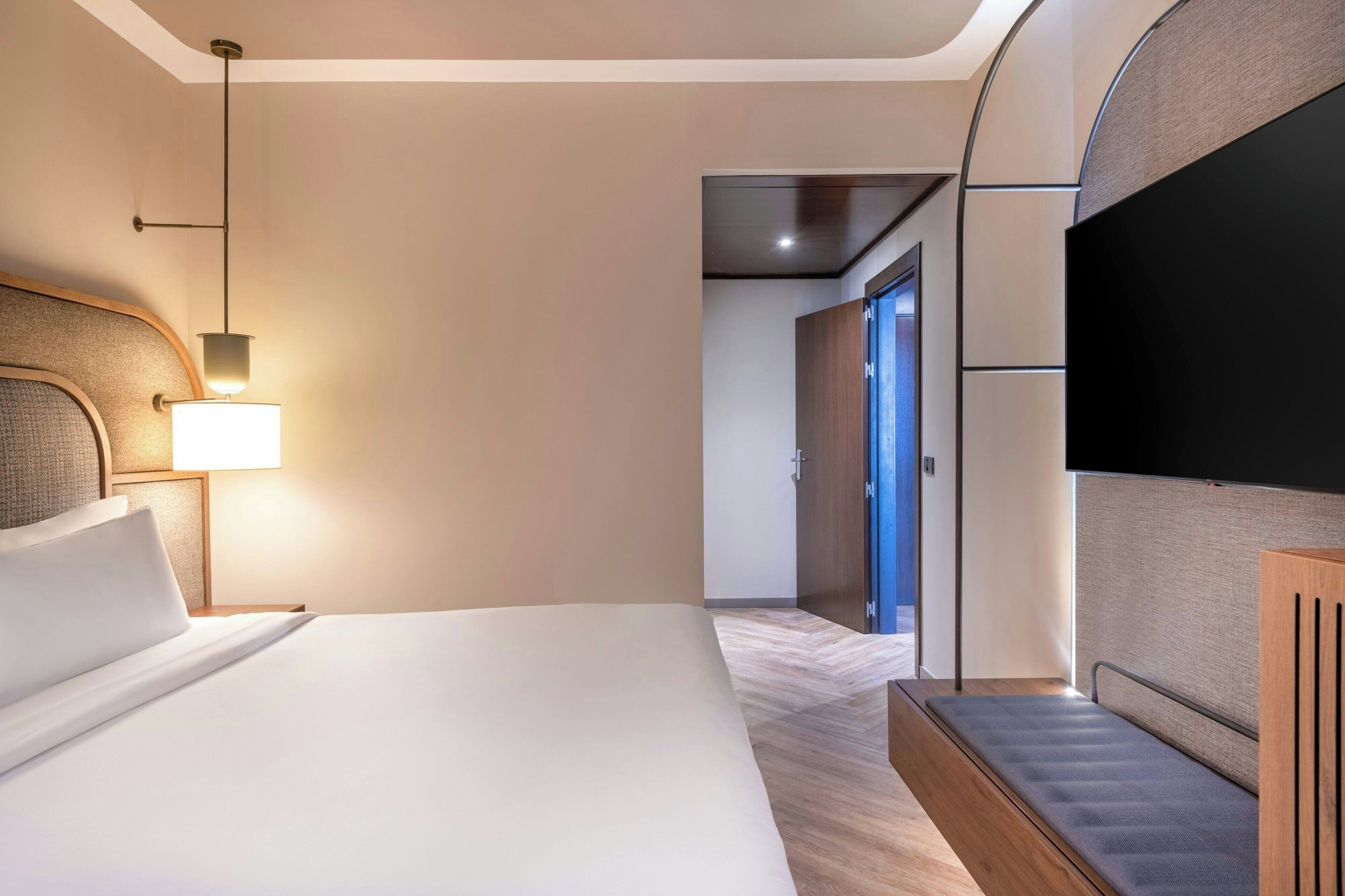 A hotel room with a bed, a TV, a lamp, and a hallway leading to a door.