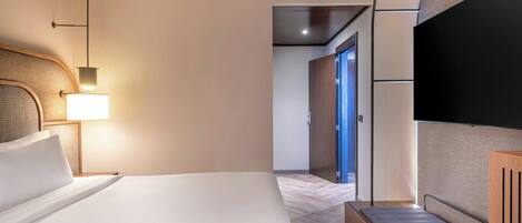 A hotel room with a bed, a TV, a lamp, and a hallway leading to a door.