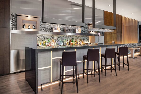 A modern bar area with a long counter, multiple stools, and a variety of bottles and glasses. - AC Hotel Palacio Universal by Marriott (Vigo)
