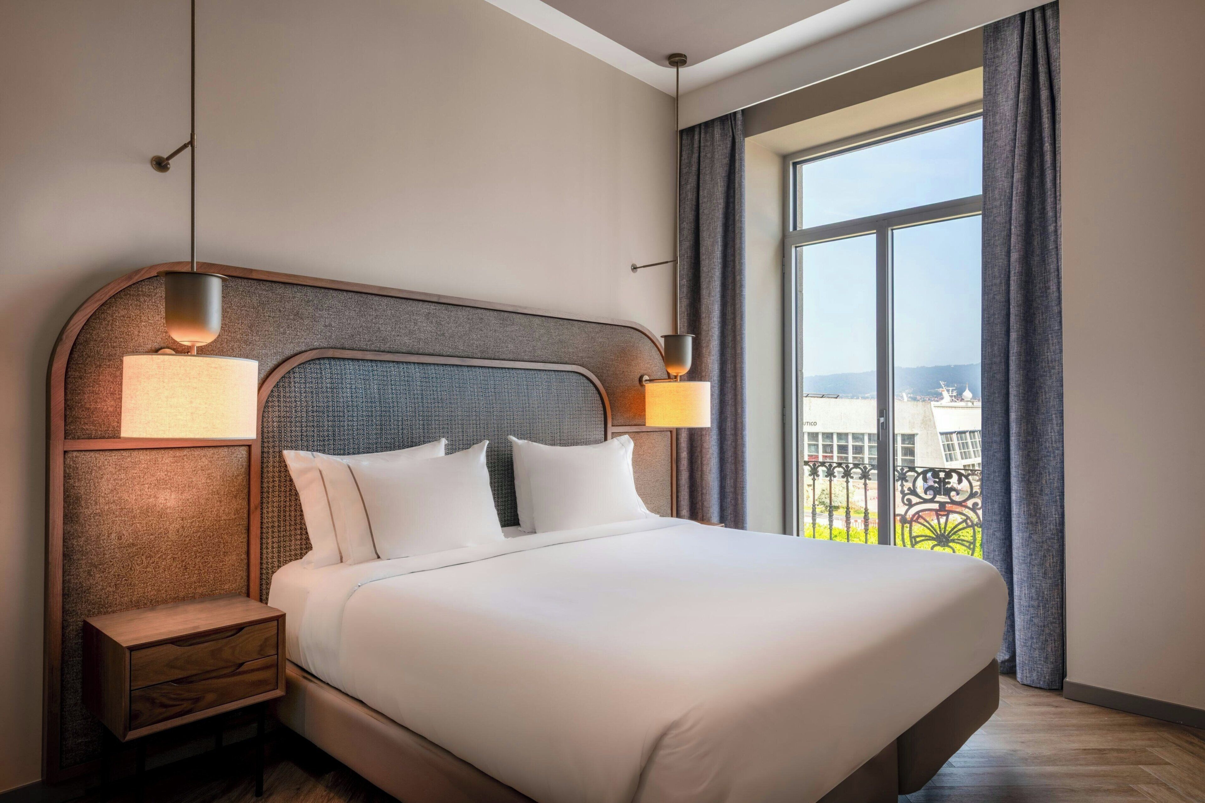 Habitación de hotel con una cama grande, dos lámparas de noche, una mesita de noche y un ventanal con vistas a edificios.