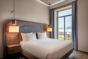 Habitación de hotel con una cama grande, dos lámparas de noche, una mesita de noche y un ventanal con vistas a edificios.