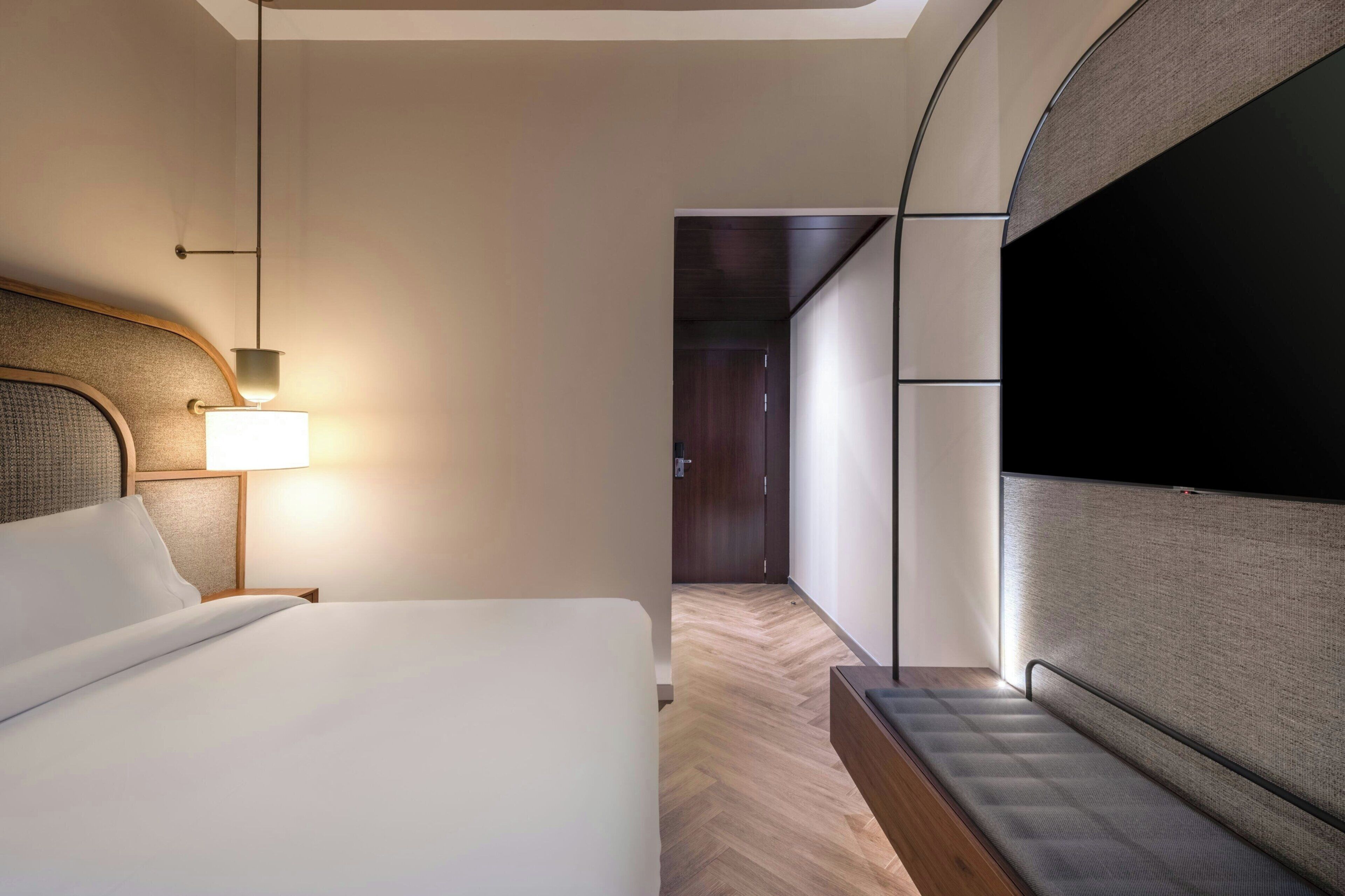 Quarto de hotel com cama, luminária de cabeceira, televisão e piso de madeira.