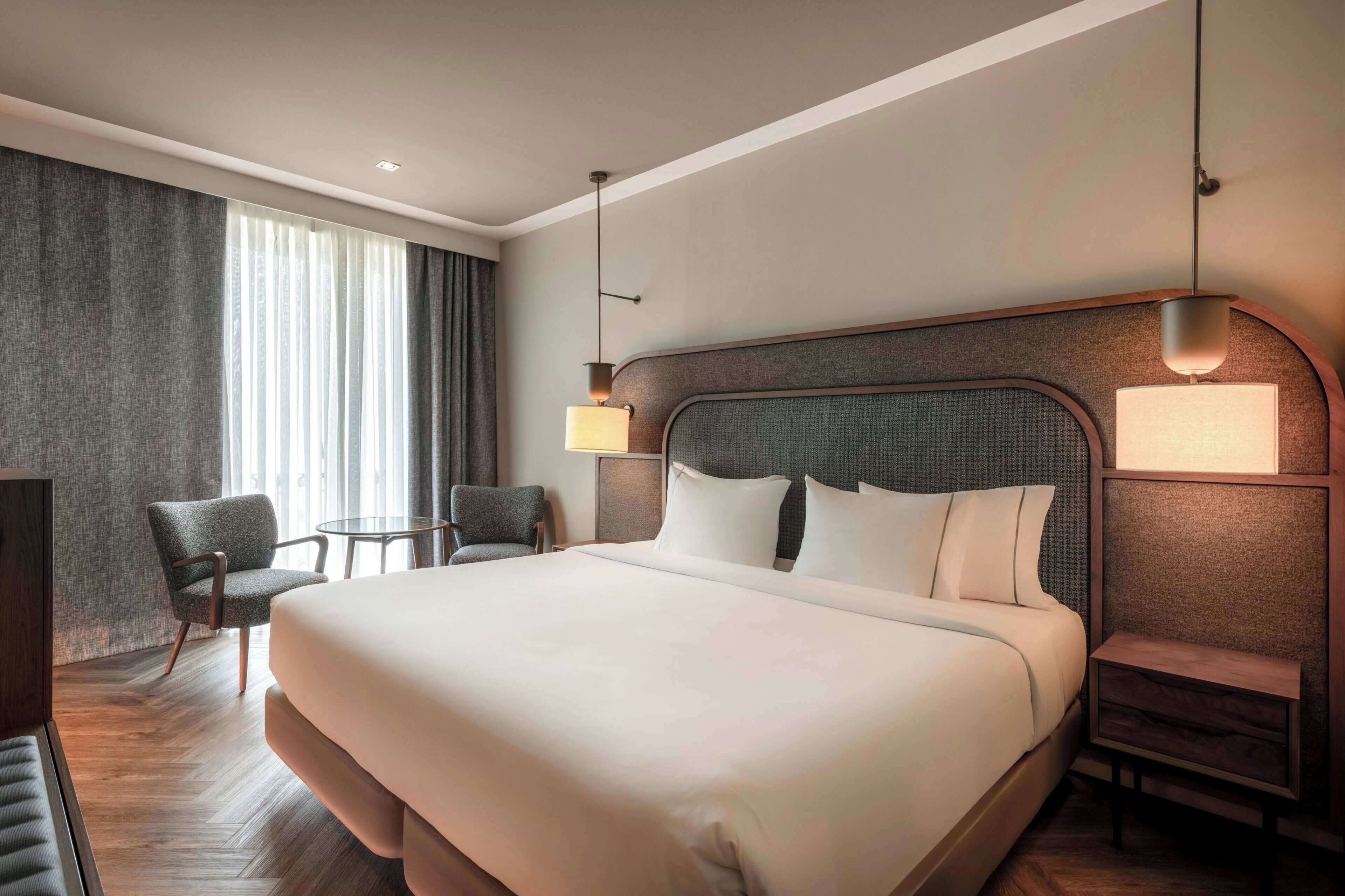 Une chambre d’hôtel avec un grand lit, deux tables de chevet, une chaise, une petite table et une grande fenêtre avec des rideaux.