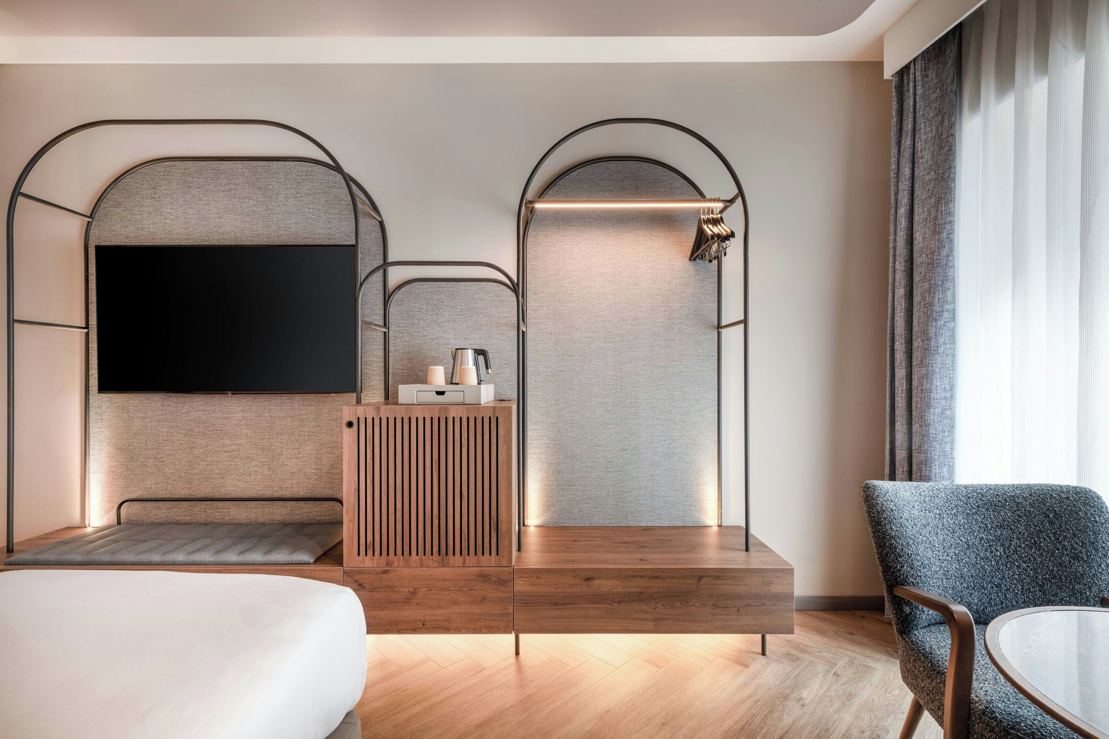 A modern hotel room with a large flat-screen TV, a wooden console table, a seating area with a sofa and armchair, and a large window with curtains.