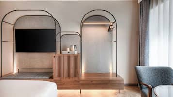 A modern hotel room with a large flat-screen TV, a wooden console table, a seating area with a sofa and armchair, and a large window with curtains.