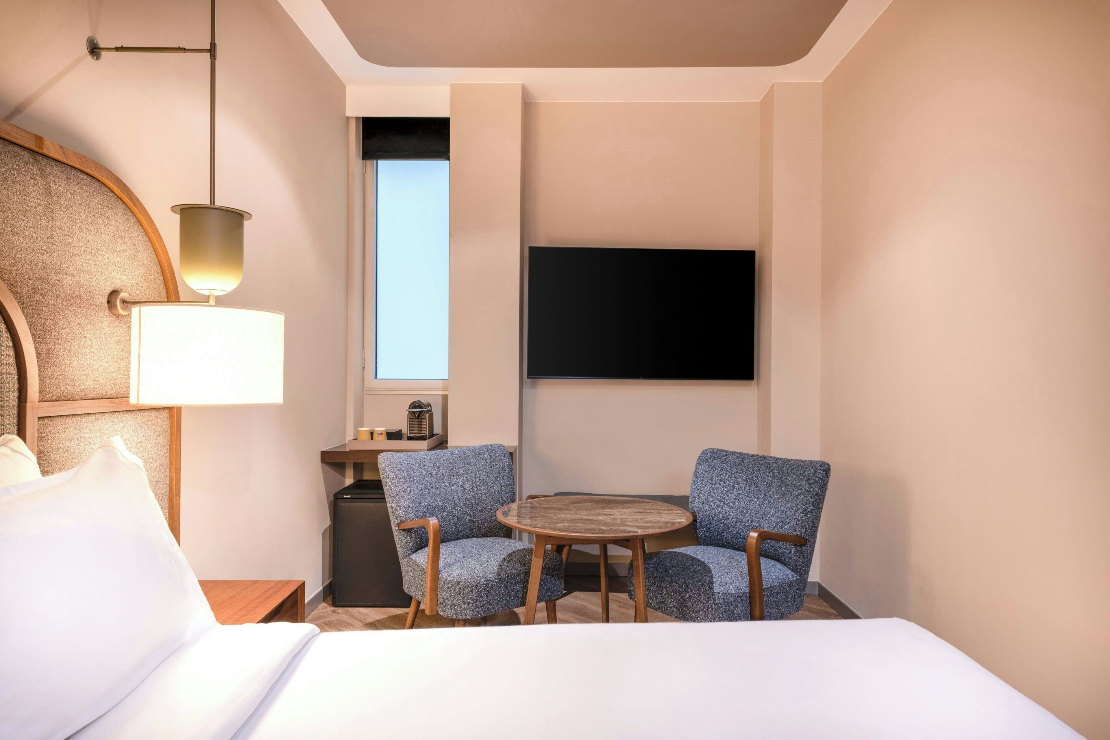 A hotel room with a bed, two armchairs, a small table, a lamp, a TV, and a window.