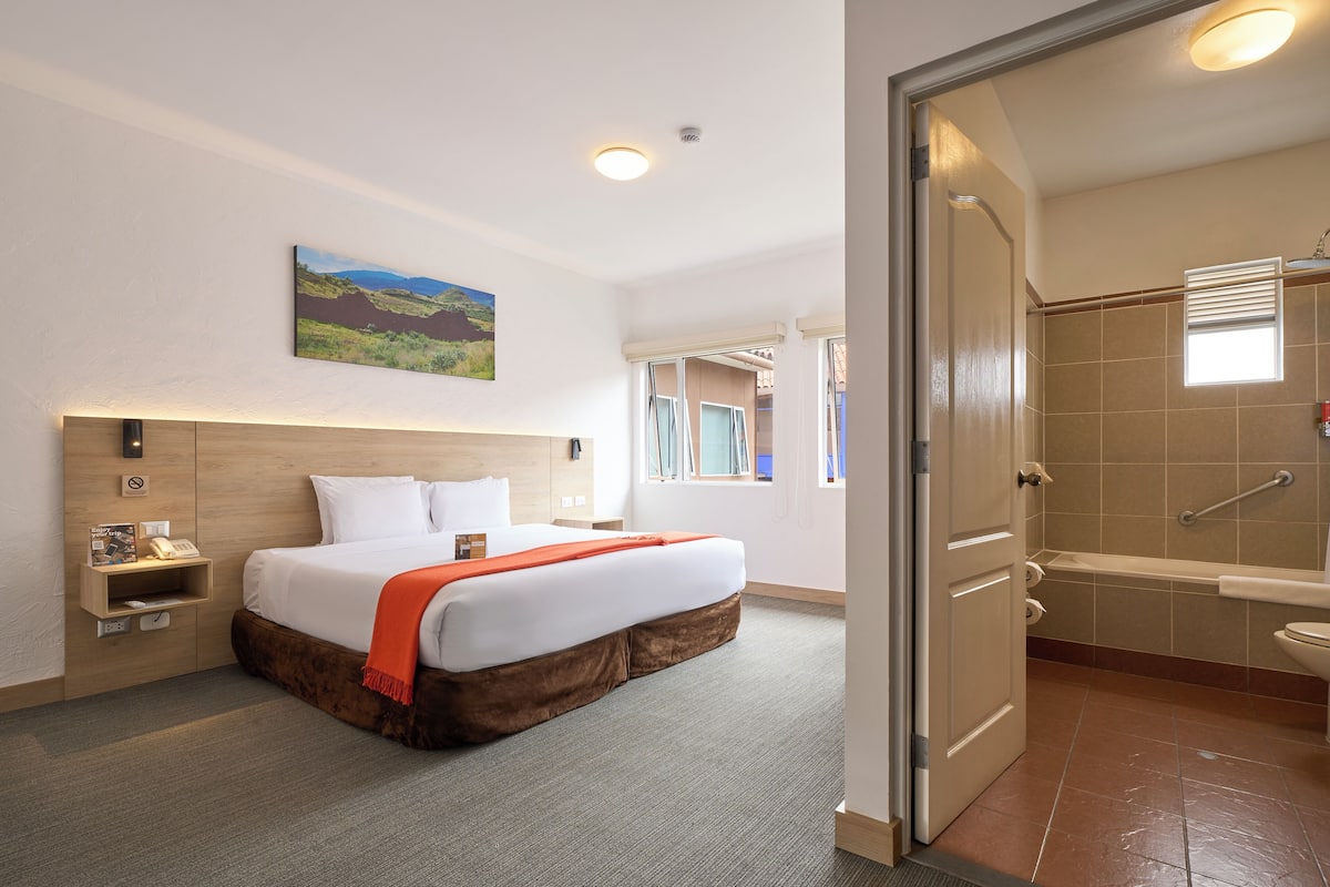 A hotel room with a bed, a nightstand, a painting, a bathroom with a shower, and a window.