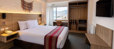 Habitación de hotel con cama, mesita de noche, lámpara, televisor, armario y vista a la ciudad.