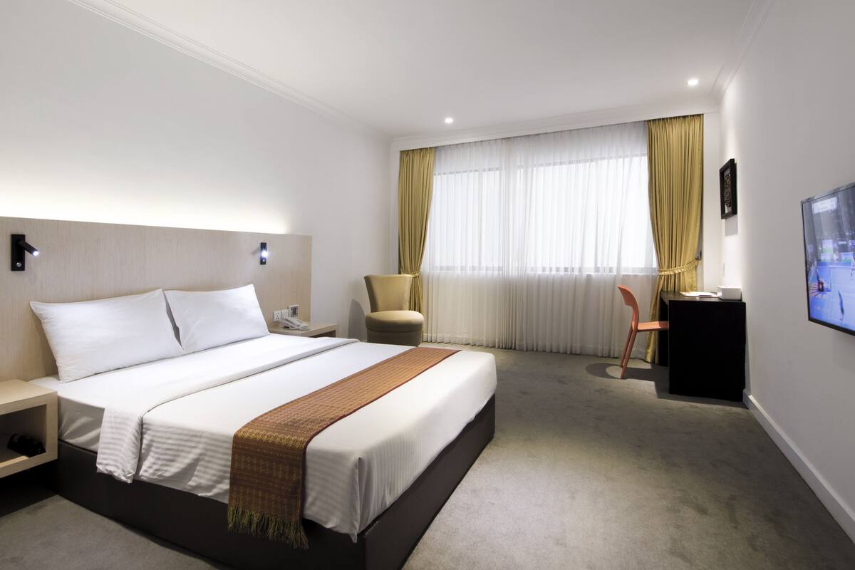 Habitación de hotel con cama, escritorio, silla, televisor y ventana con cortinas.