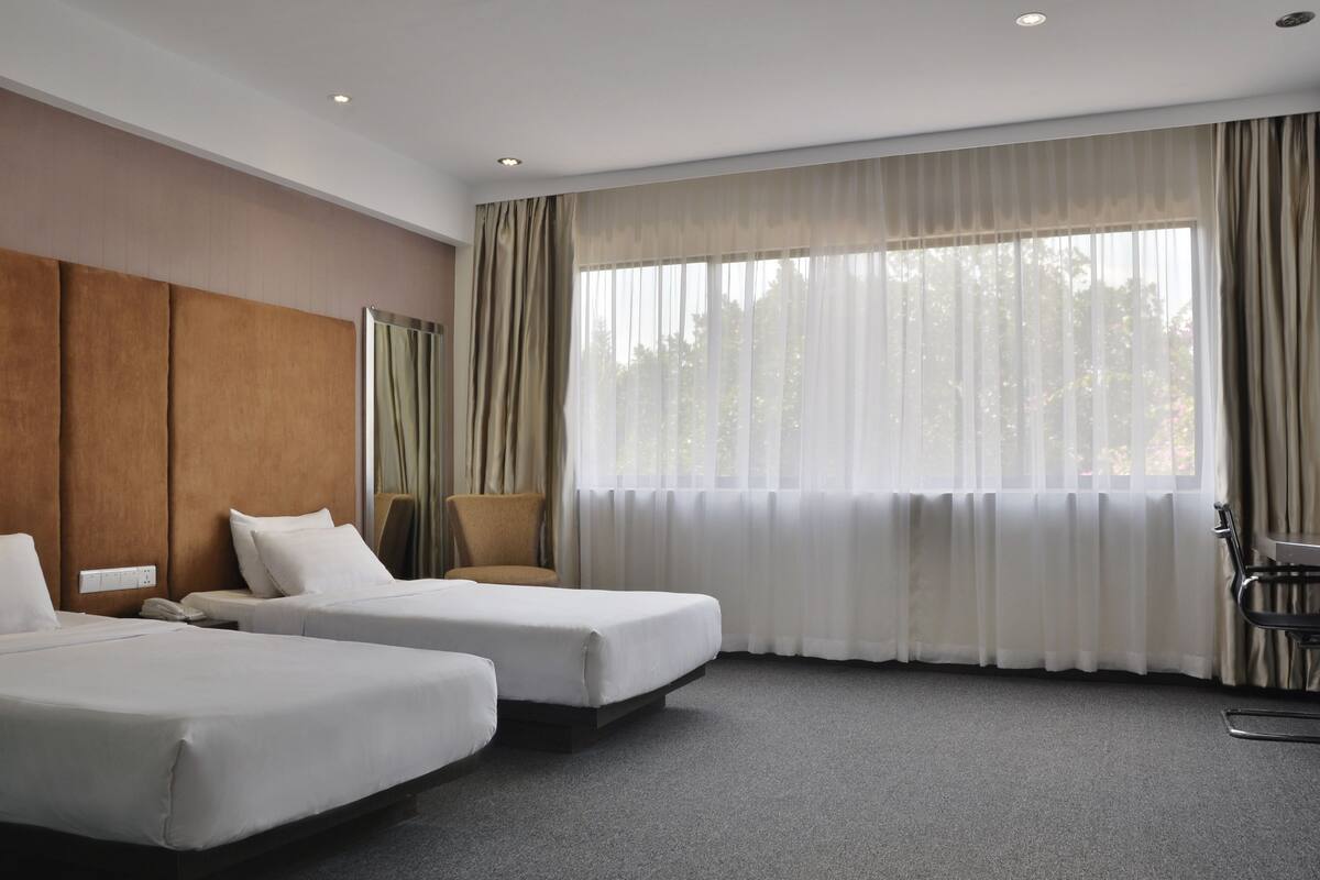 Ein Hotelzimmer mit zwei Betten, einem großen Fenster mit durchsichtigen Vorhängen, einem Holz-Kopfteil und einem grauen Teppich.