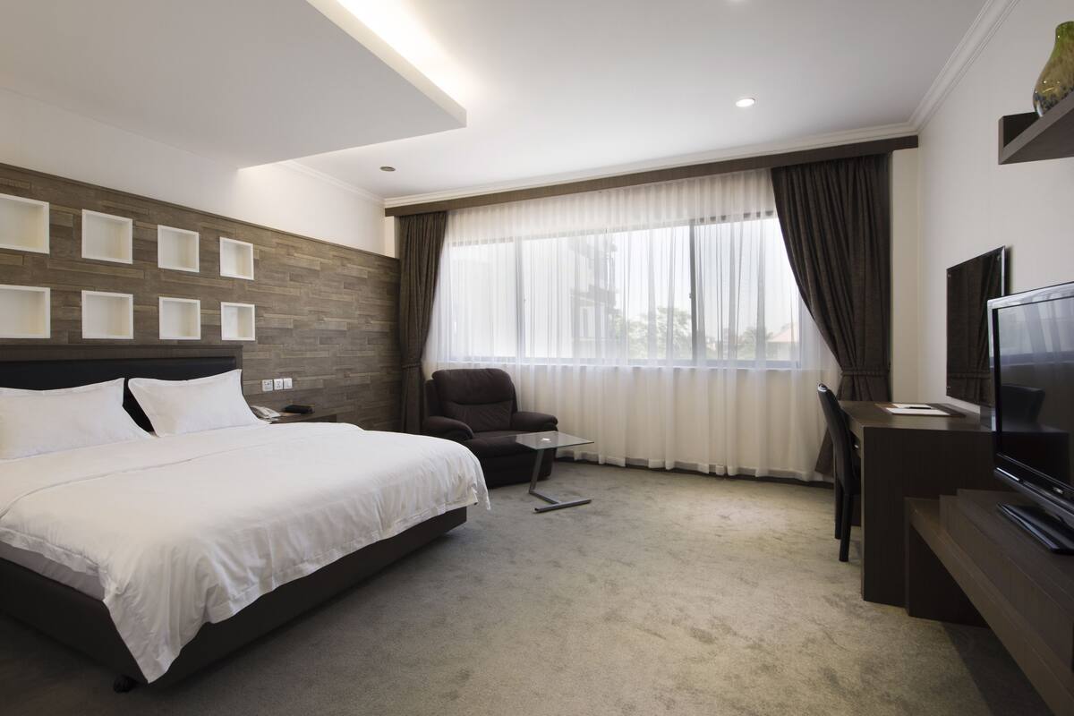 Una habitación de hotel moderna con una cama grande, un escritorio con televisor, una silla y una ventana con cortinas.