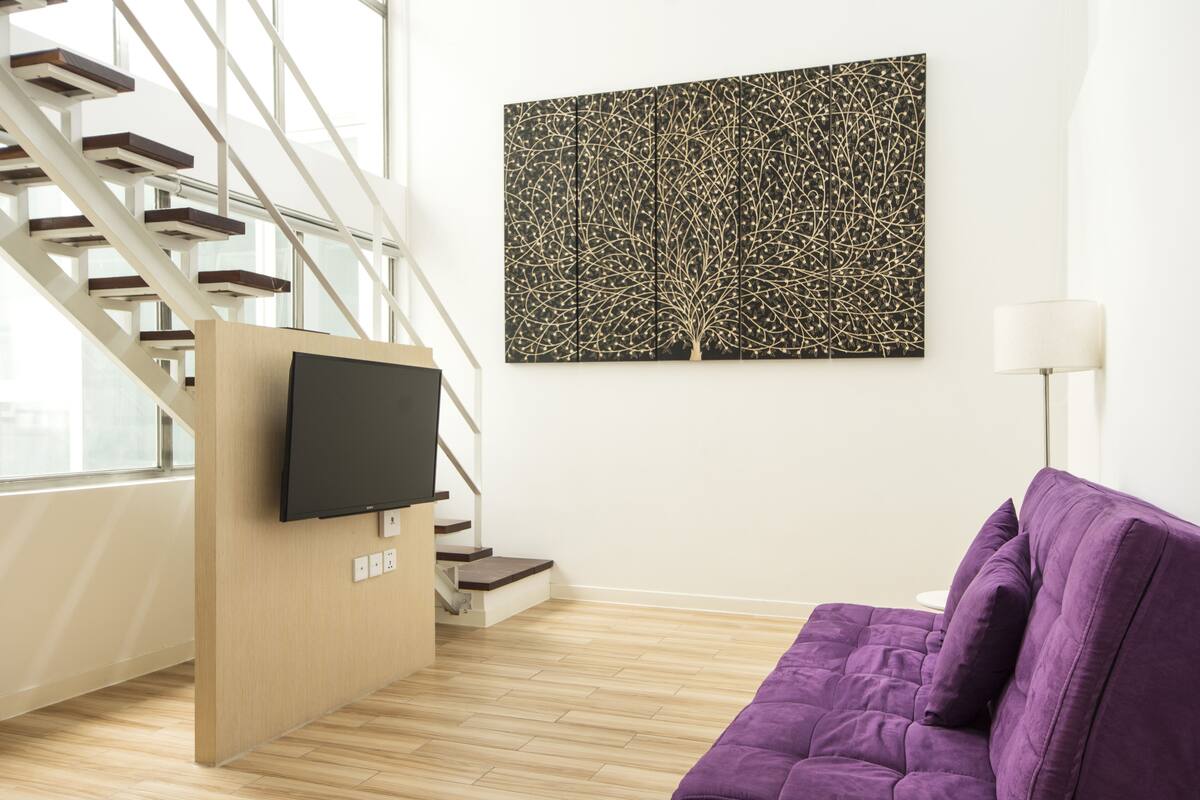 Ein modernes Wohnzimmer mit einem violetten Sofa, einem flachbildfernseher, der an einer Holzverkleidung angebracht ist, einer Treppe mit Glasgeländern und einem abstraktem Kunstwerk an der Wand.