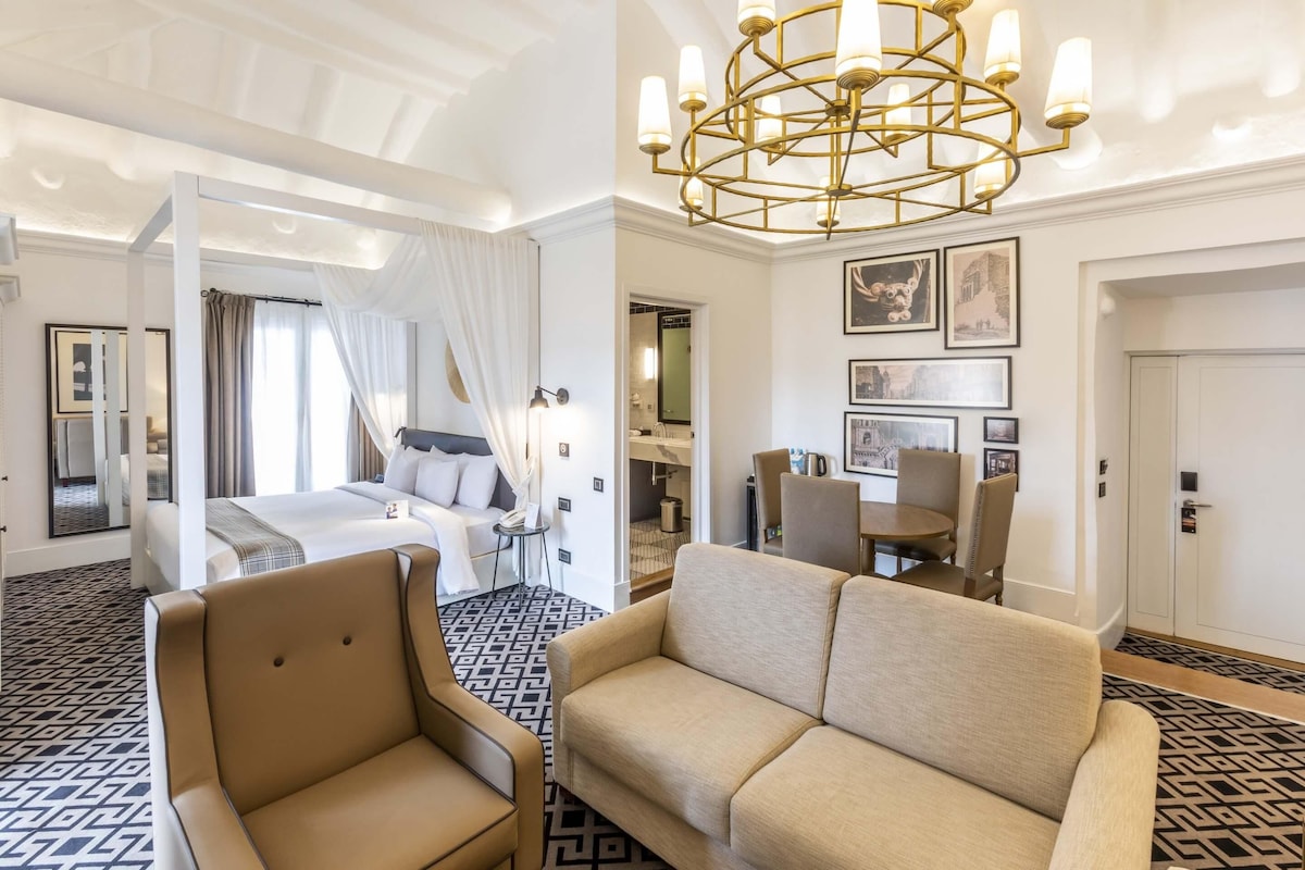 A hotel room with a bed, a sitting area with a sofa and armchair, a dining table with chairs, and a chandelier.