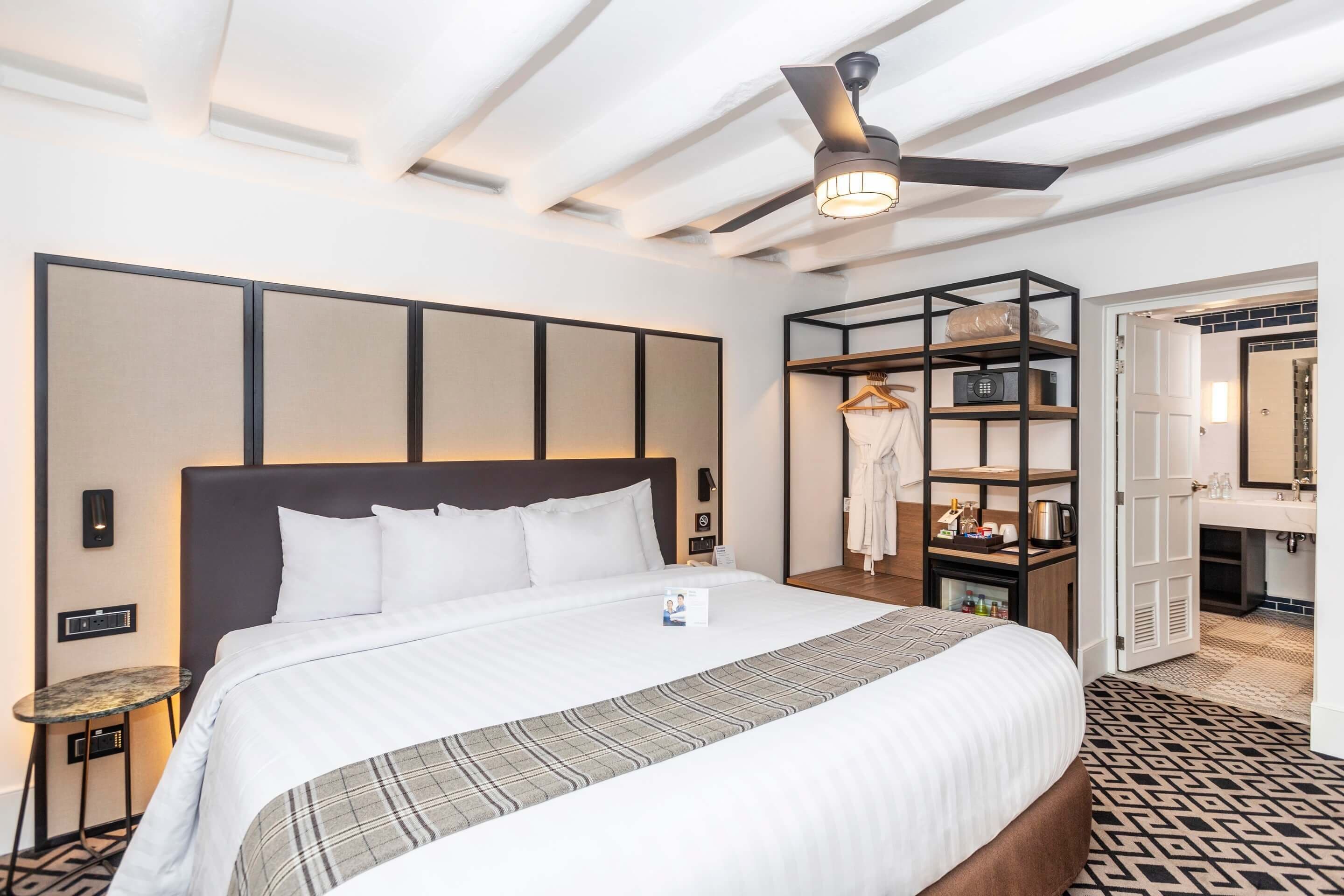 A hotel room with a large bed, a ceiling fan, a small table, a wardrobe, and a bathroom with a mirror and sink.