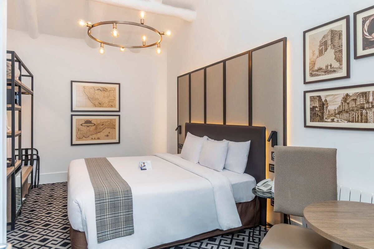 A hotel room with a bed, a desk, a chair, a map on the wall, and a light fixture.