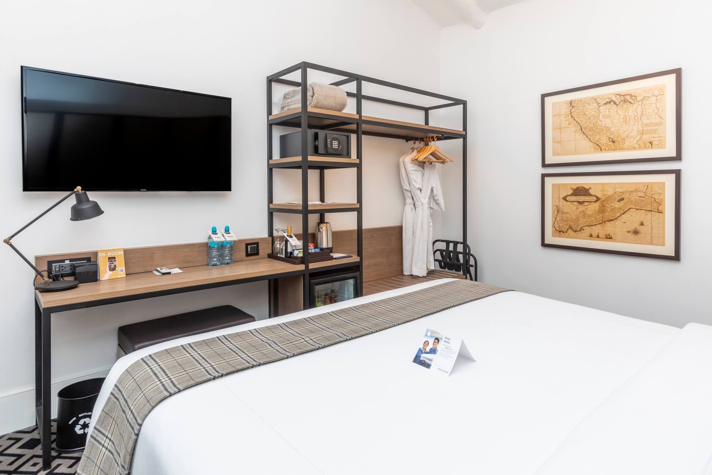 Een hotelkamer met een bed, een bureau met een computer, een televisie, een kledingkast en twee ingelijste kaarten aan de muur.