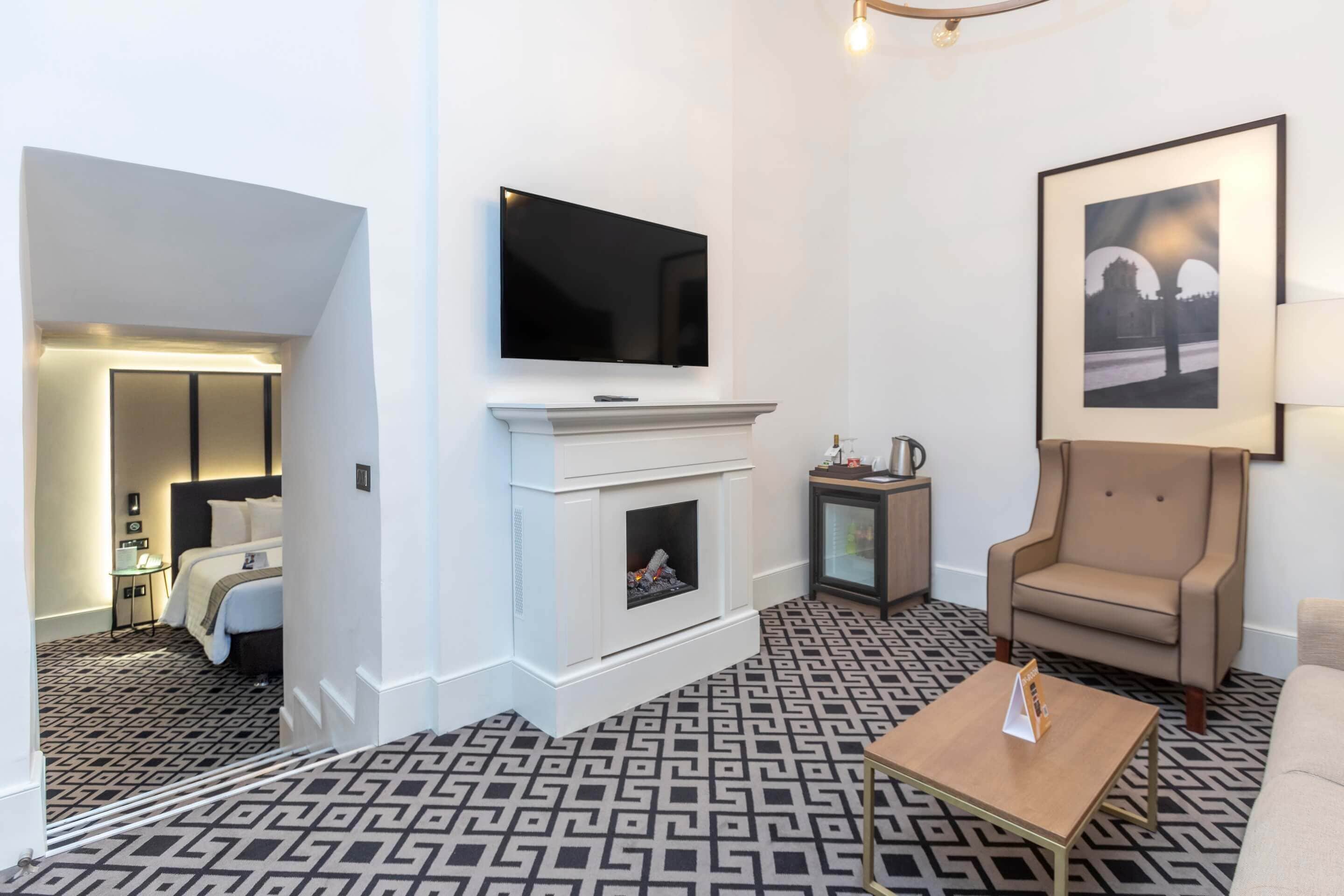A modern hotel room with a fireplace, a flat-screen TV, a framed picture, a sofa, and a patterned carpet.