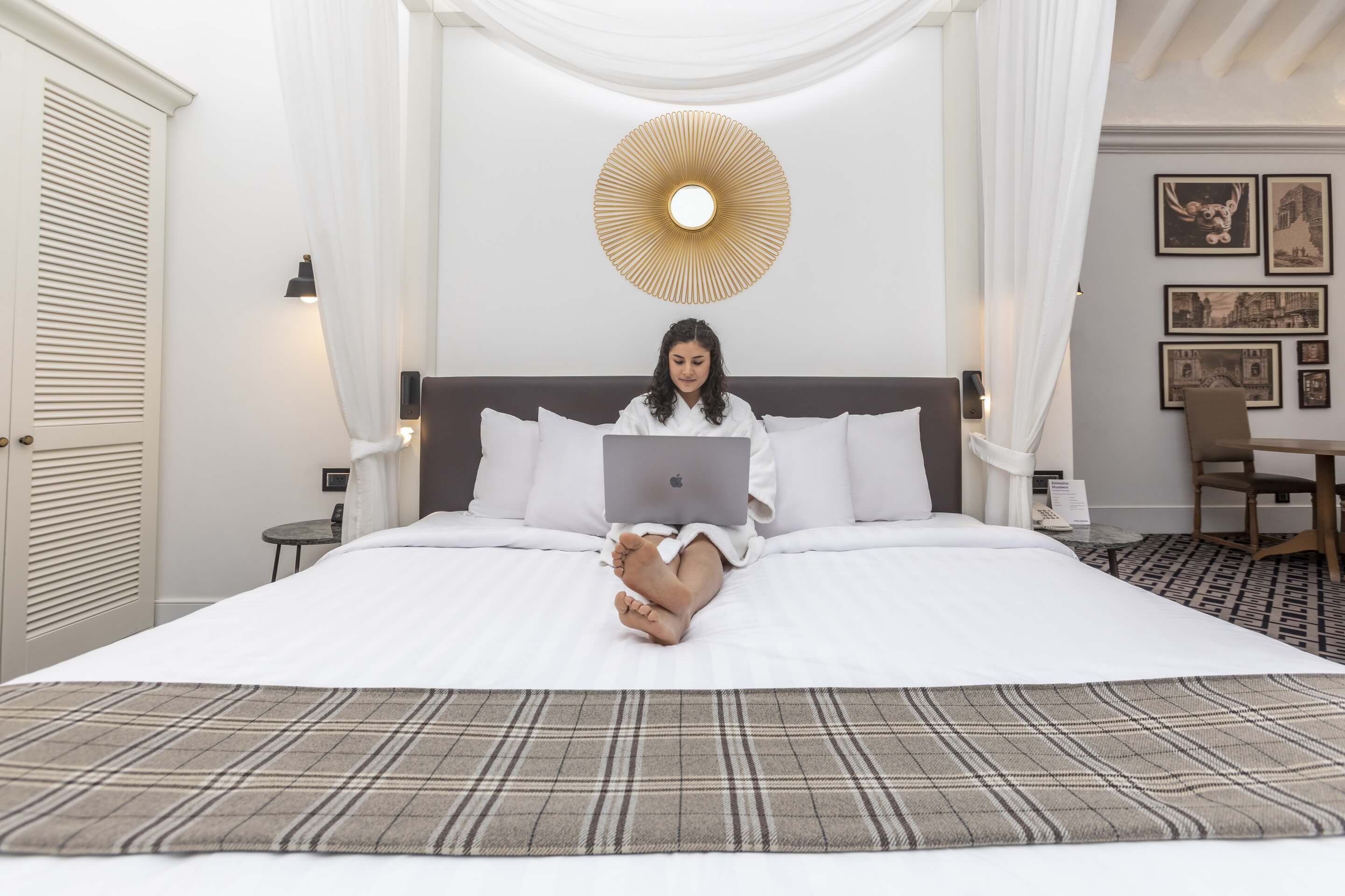 A bed with a laptop, white bedding, a plaid throw, and a wall-mounted light fixture.
