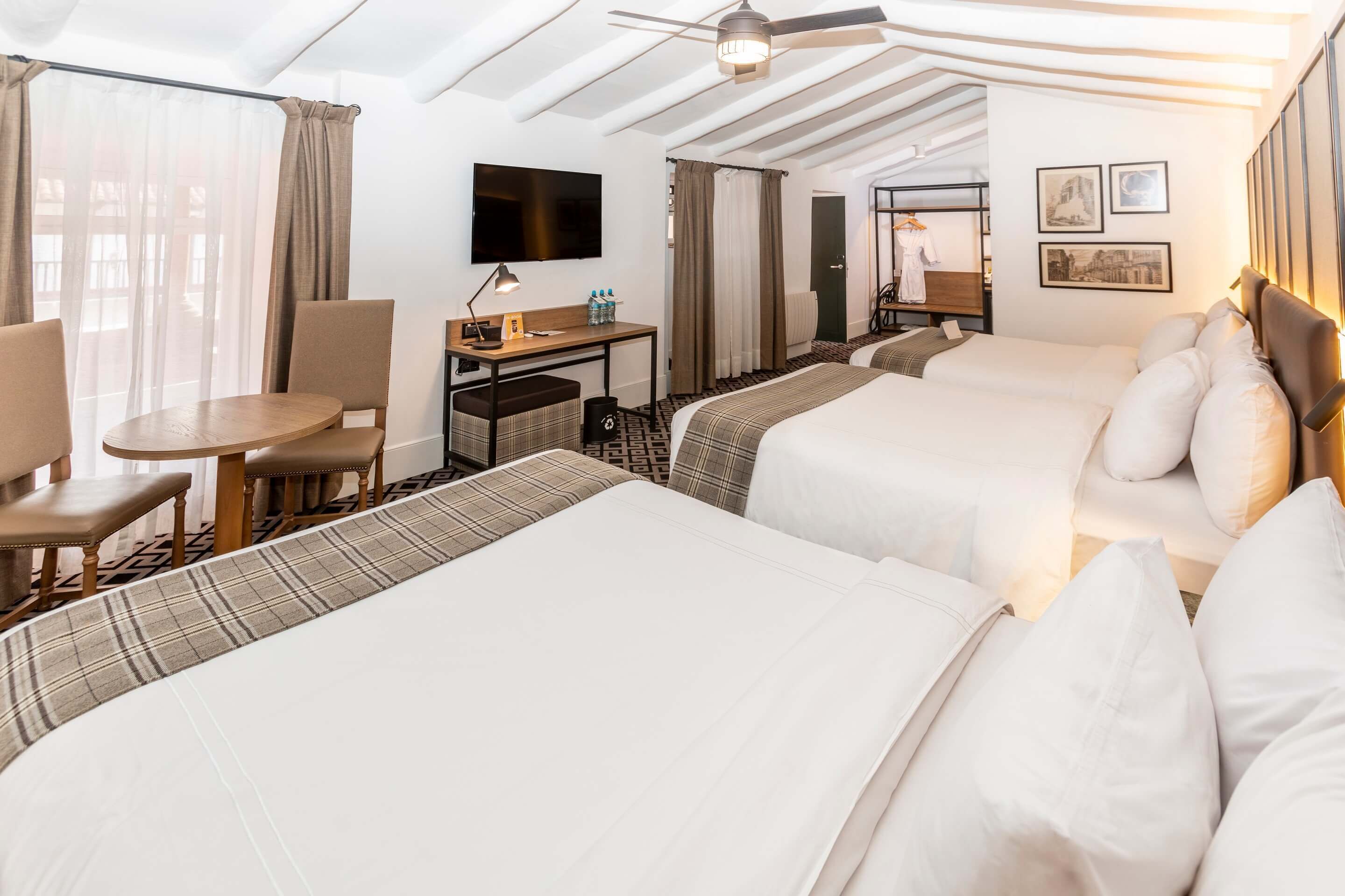 A hotel room with two beds, a desk, a chair, a TV, and a ceiling fan.