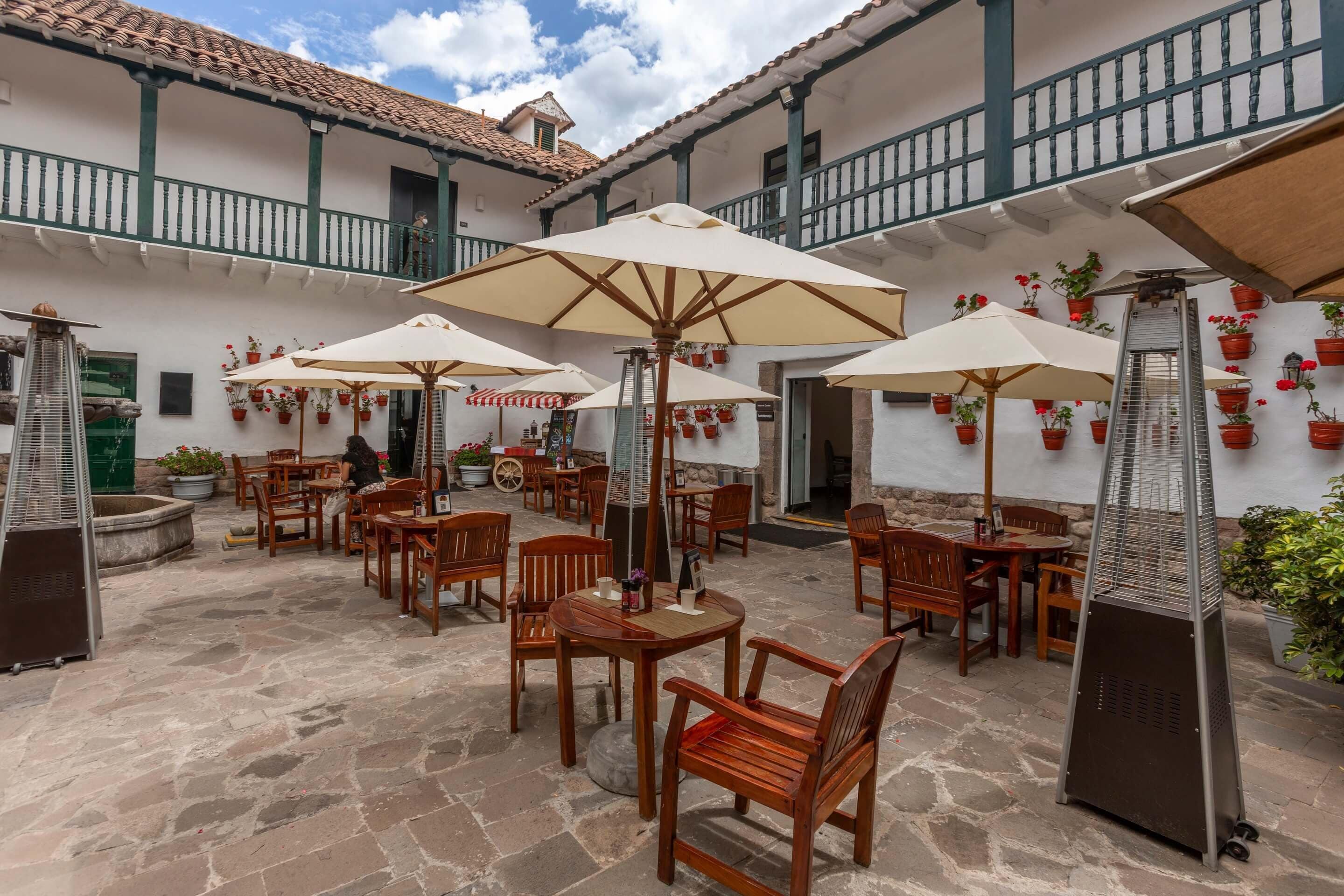 An outdoor patio with wooden tables and chairs, surrounded by potted plants and umbrellas.