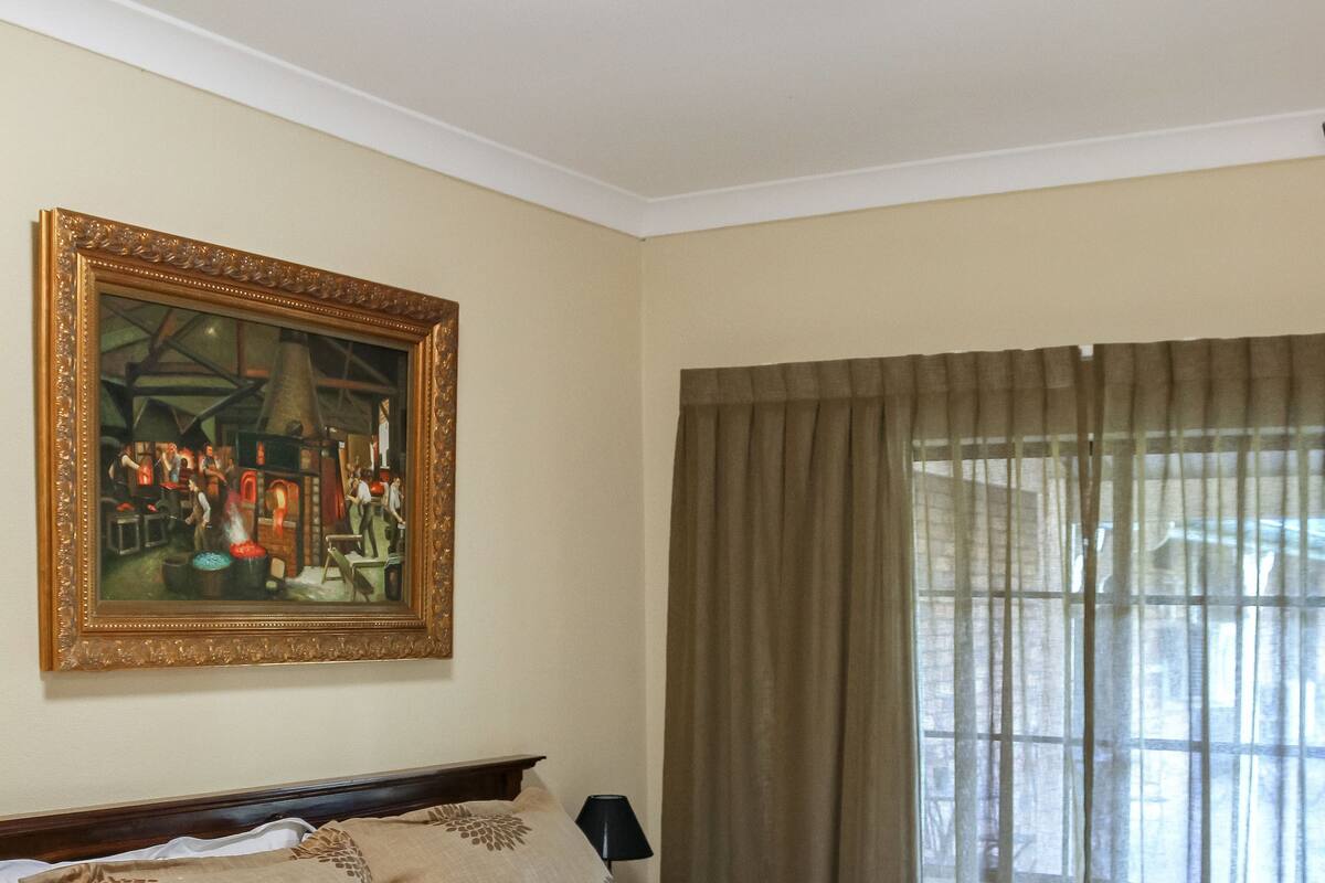 A neatly made bed with a white and beige bedspread, a framed painting above it, and a window with curtains.