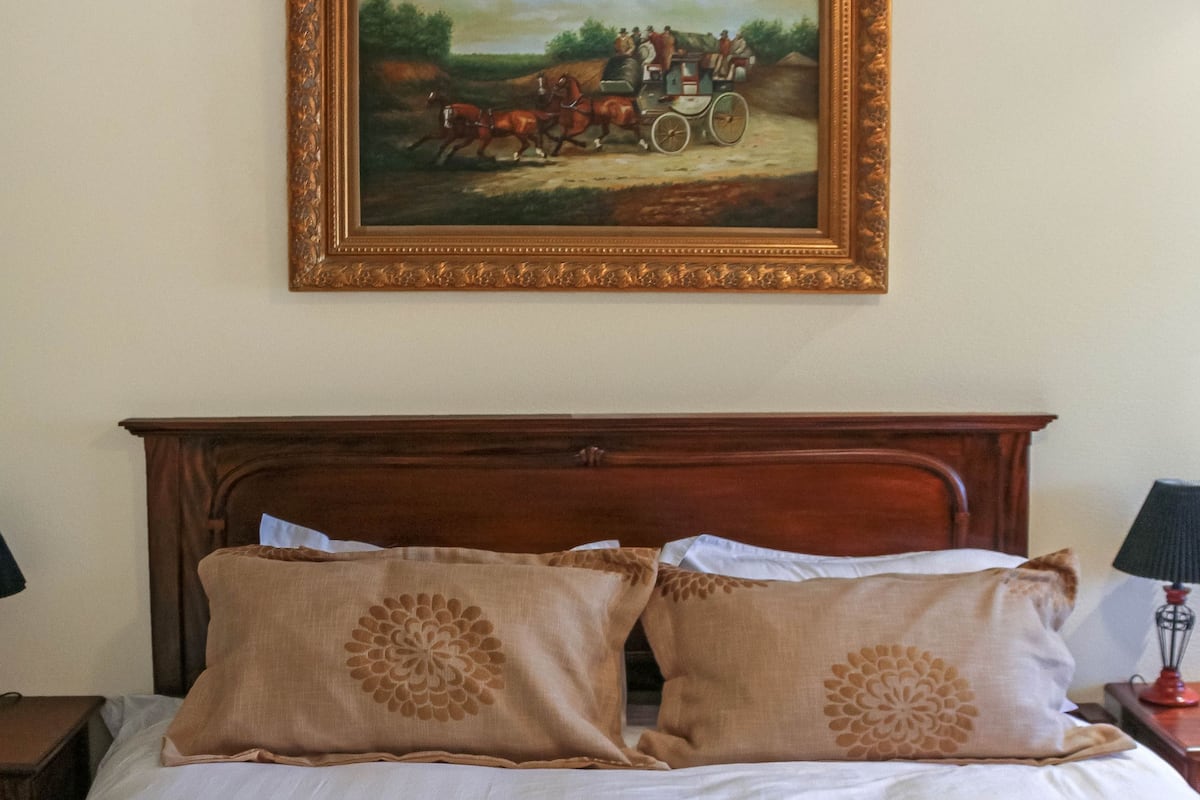 A neatly made bed with a floral pillow, a folded towel, and a painting of a horse-drawn carriage on the wall.