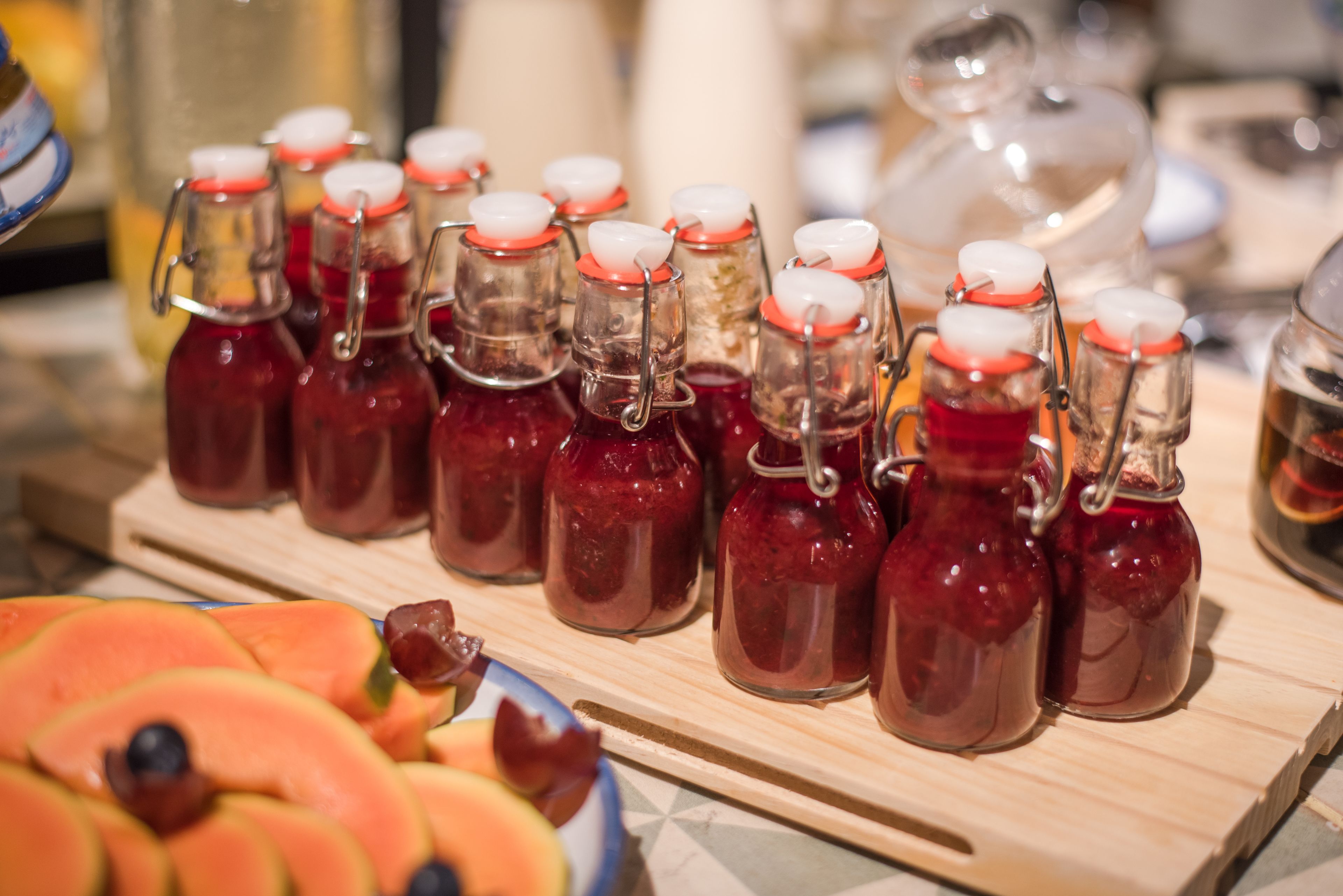 Una mesa de madera con varias botellas de vidrio que contienen un líquido rojo, presumiblemente una bebida, y un cuenco con frutas cortadas en primer plano.