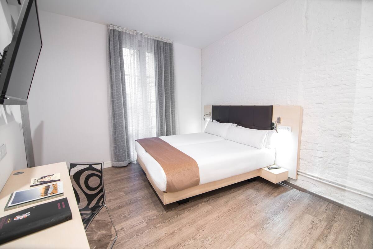 Habitación de hotel con cama, mesita de noche, televisor y ventana con cortinas.