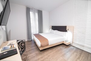 Quarto de hotel com cama, mesa de cabeceira, televisão e janela com cortinas.