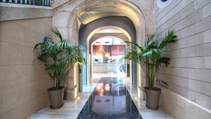 Un pasillo con piso negro pulido, flanqueado por paredes color beige y plantas en macetas. Un arco de entrada da a una recepción.