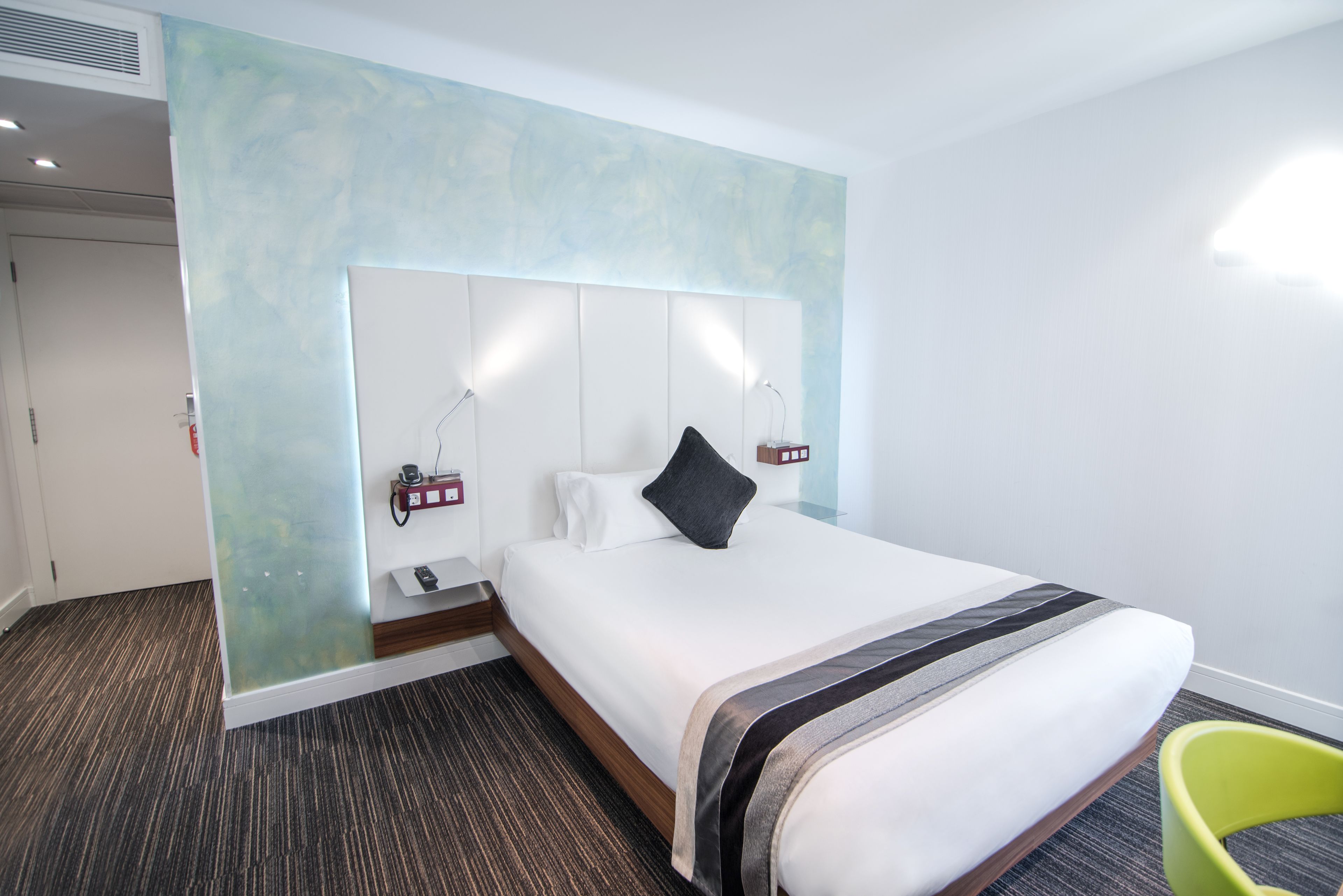 A hotel room with a large bed, a headboard with integrated lighting, a bedside table with a lamp, and a green chair.