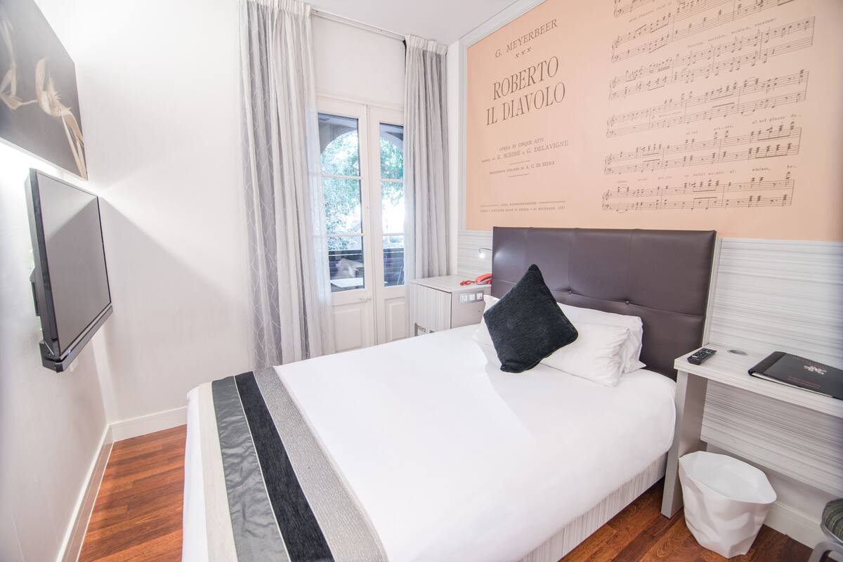 Habitación de hotel con una cama grande, un televisor de pantalla plana montado en la pared, una ventana con cortinas y una pared con partituras.