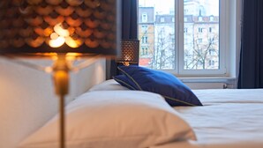 A bed with white bedding and a blue pillow, a lamp with a patterned shade, and a window with a view of buildings. - Saint Georges Hotel (Zürich)