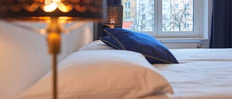 Una cama con ropa de cama blanca y una almohada azul, una lámpara con pantalla estampada, y una ventana que ofrece vistas a edificios.
