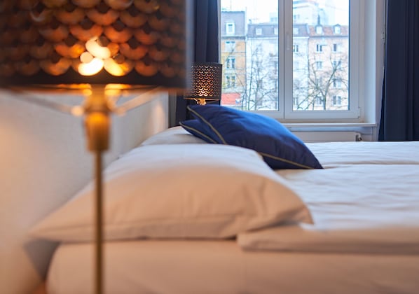 A bed with white bedding and a blue pillow, a lamp with a patterned shade, and a window with a view of buildings. - Saint Georges Hotel (Zürich)