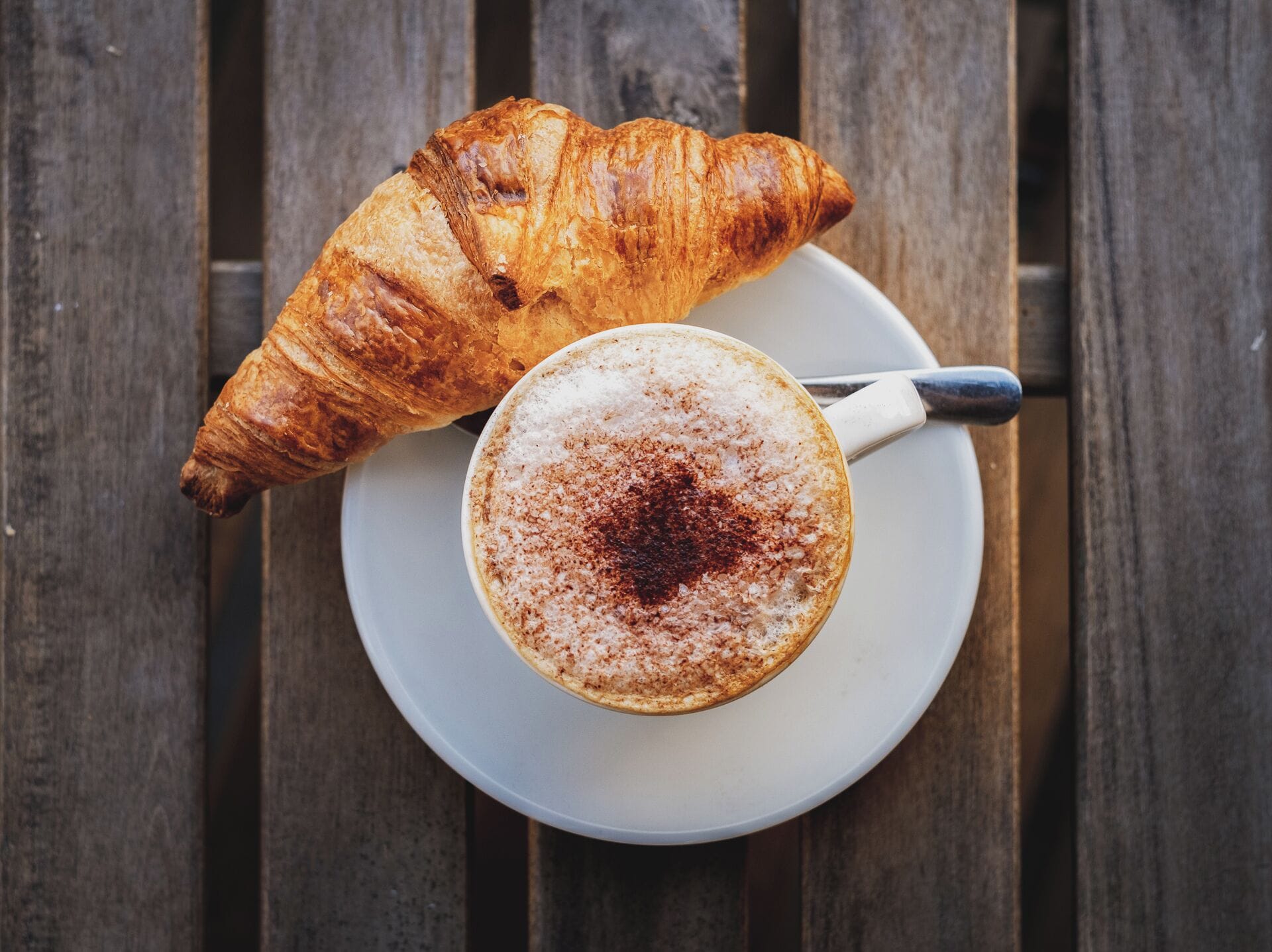 Ein Croissant und eine Tasse Kaffee auf einem weißen Teller auf einer Holztischplatte.