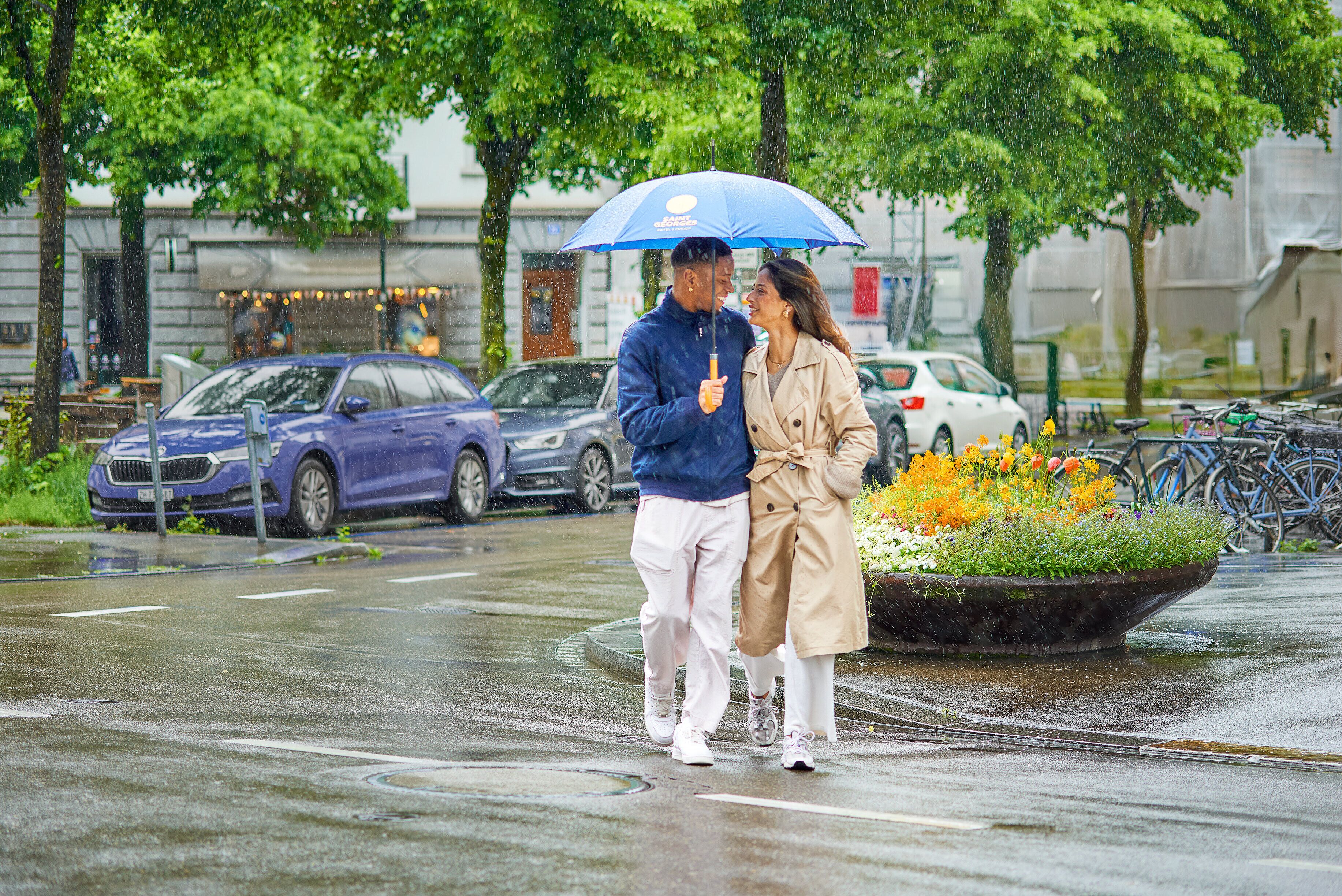 Una pareja caminando sobre una calle mojada con autos estacionados al lado, un cantero con diversas flores y bicicletas aparcadas al fondo.