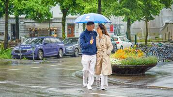 Una pareja caminando sobre una calle mojada con autos estacionados al lado, un cantero con diversas flores y bicicletas aparcadas al fondo.