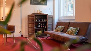 A cozy living room with a brown leather sofa, a glass coffee table, and a bookshelf filled with bottles. A chalkboard menu is visible on the wall, and a window provides a view of the outside. - Saint Georges Hotel (Zürich)