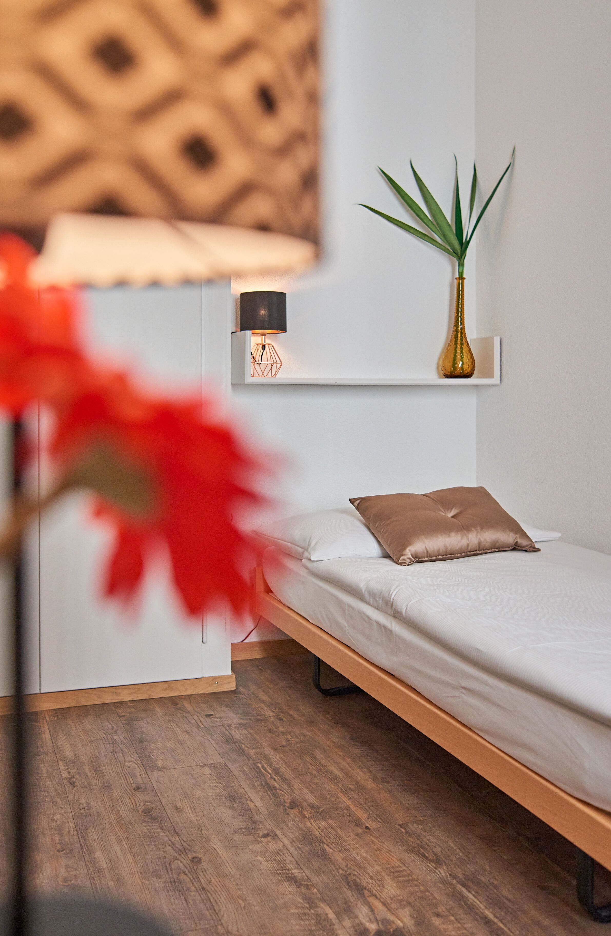 A minimalist bedroom with a wooden bed, a bedside table with a lamp, and a potted plant on a shelf.