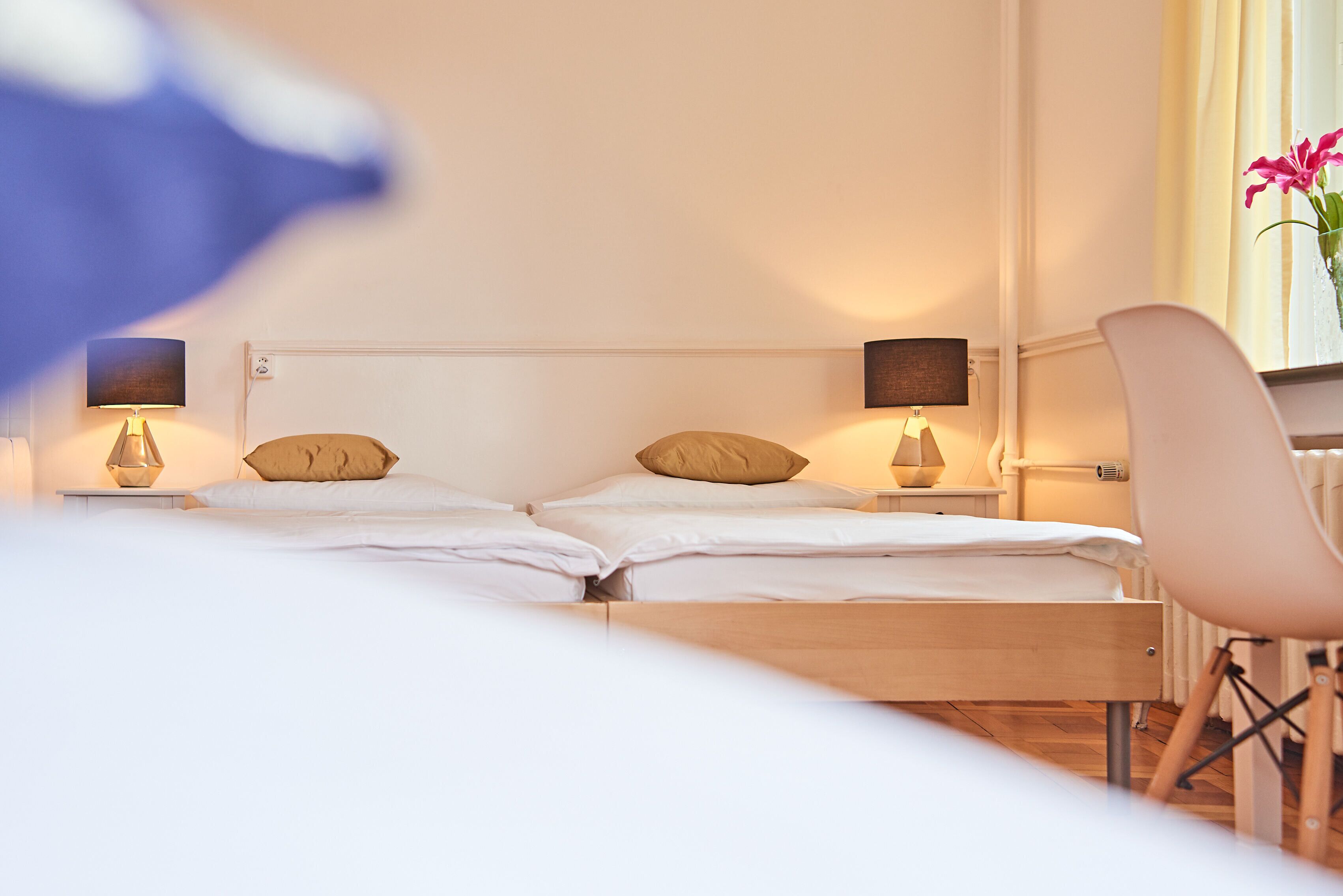 A hotel room with two beds, a wooden headboard, bedside lamps, a chair, and a window with curtains.