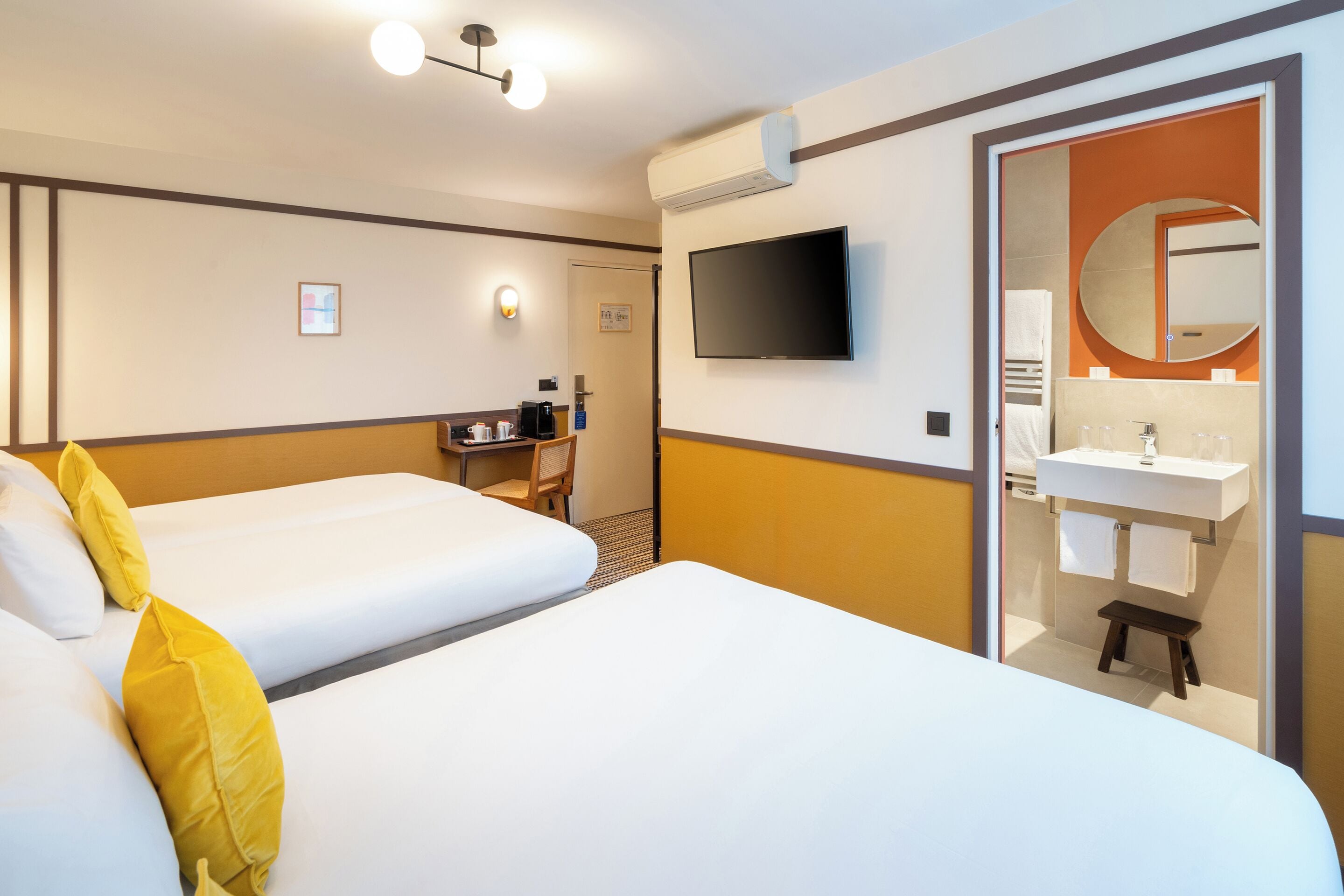 Een hotelkamer met twee bedden, een bureau, een televisie, een badkamer met wastafel en spiegel, en een klein krukje.